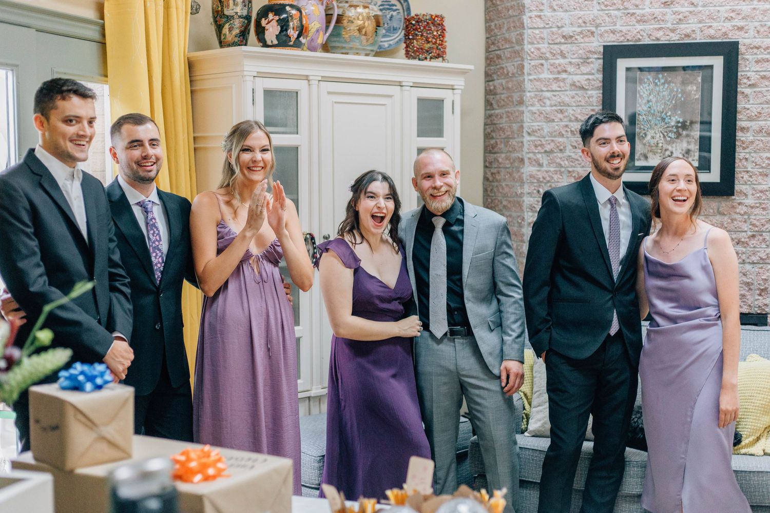 Wedding party group laughing together in living room wearing purple bridesmaid dresses and suits.