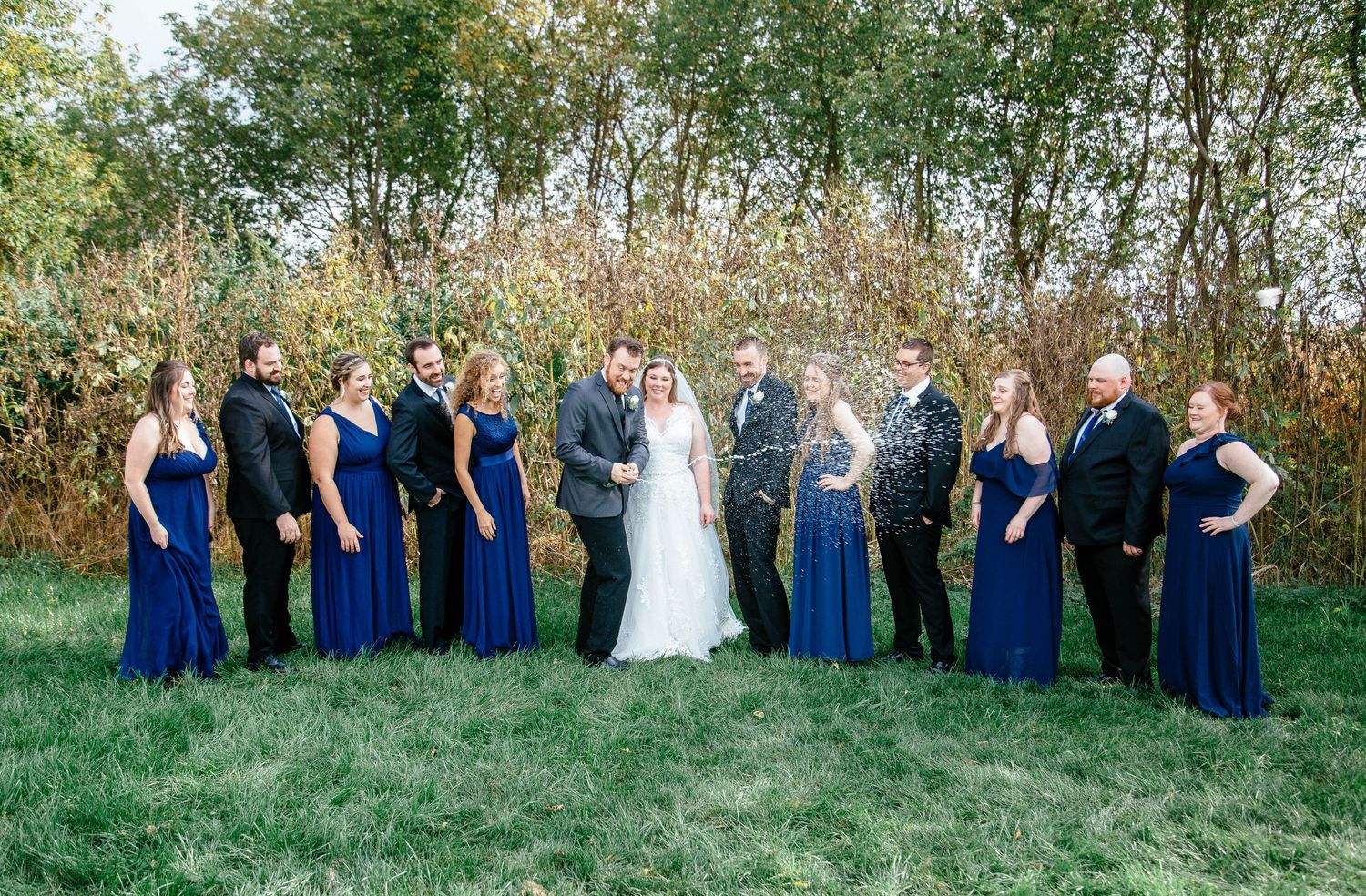 Full wedding party in royal blue and black attire celebrating with champagne on grassy outdoor venue.
