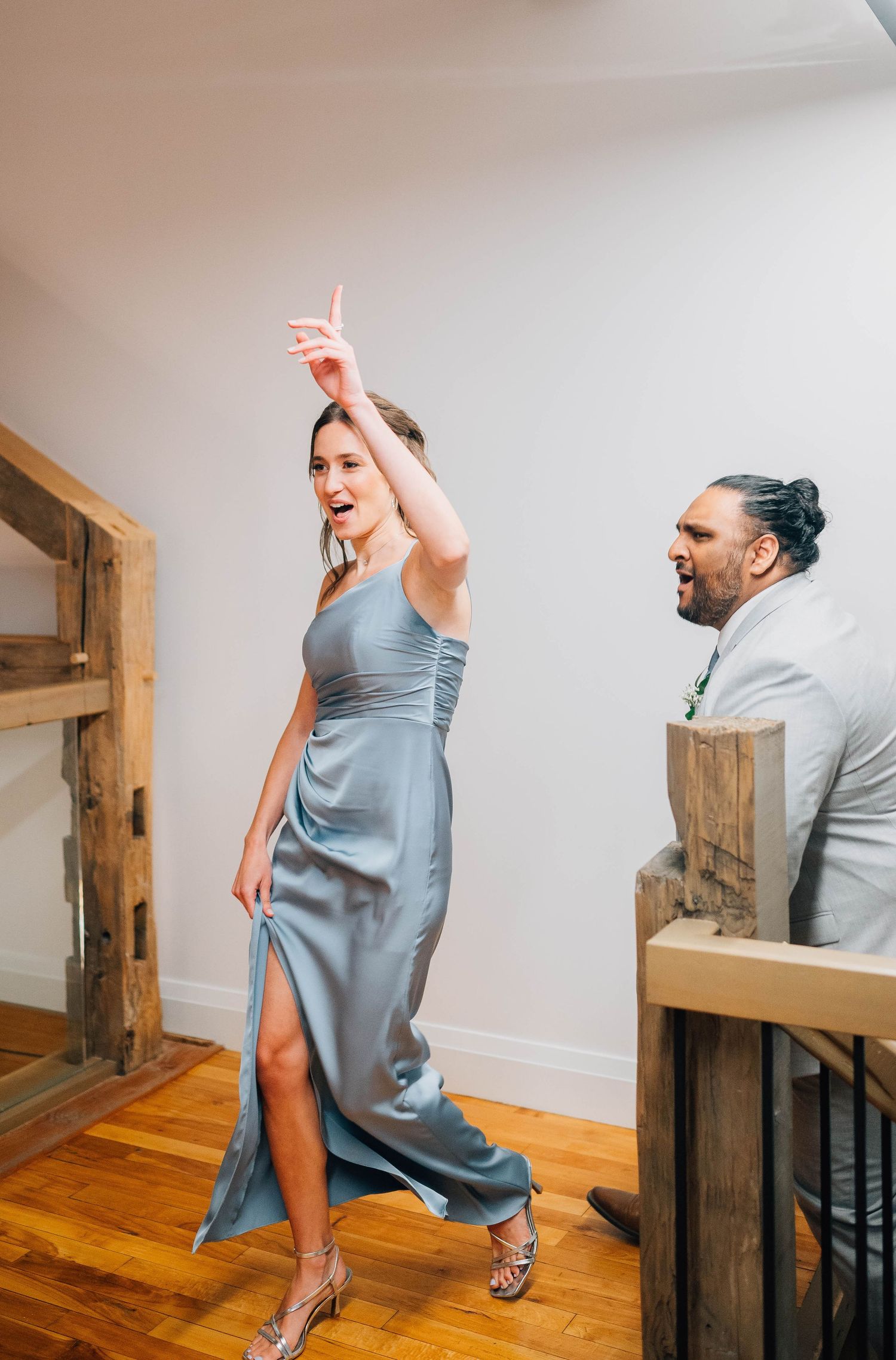 Bridesmaid in blue satin dress dancing with raised arm while groomsman watches at wedding reception.