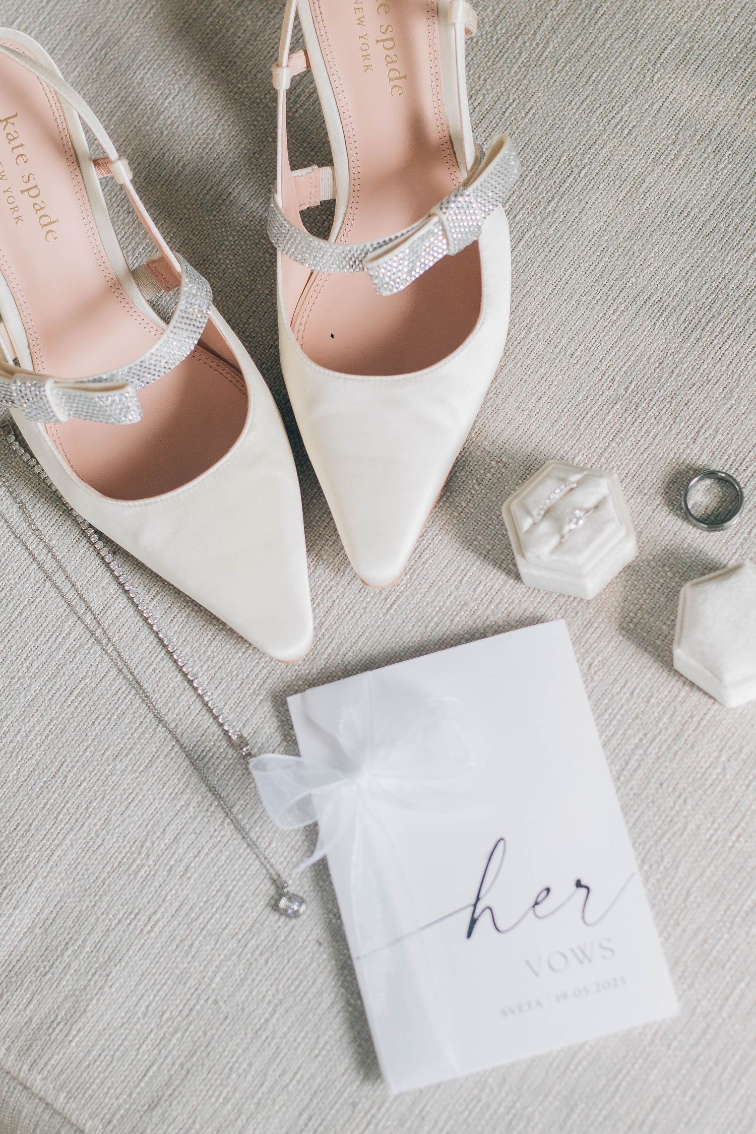 Flat lay of white Kate Spade bridal heels, vow book, necklace, and ring box on fabric.