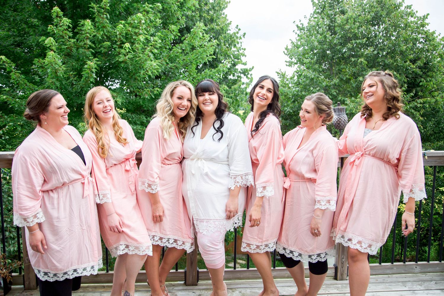 Bride in white robe laughs with six bridesmaids in matching pink lace robes on outdoor deck.