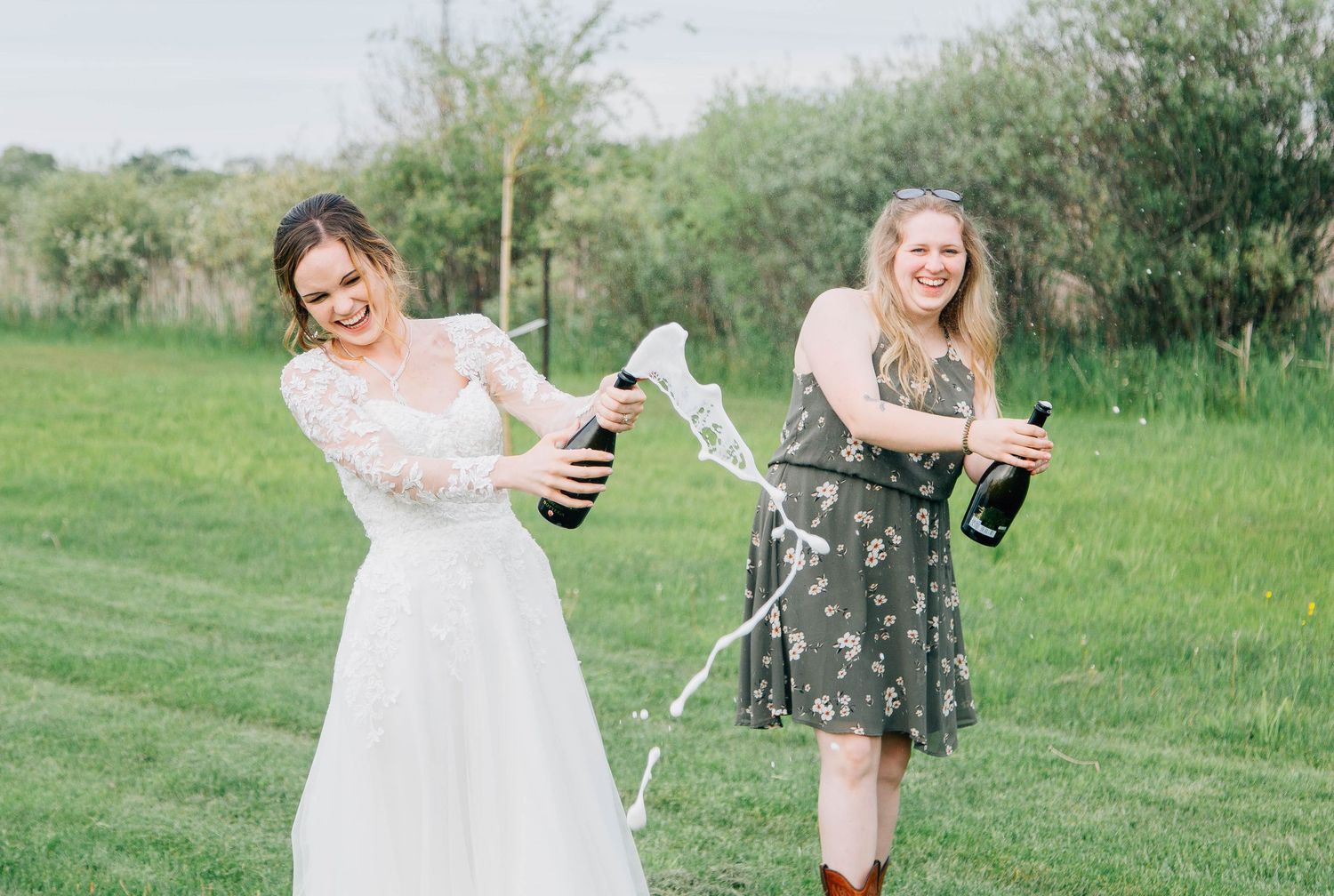 Laughing bride in lace gown and bridesmaid spraying champagne on a green lawn.