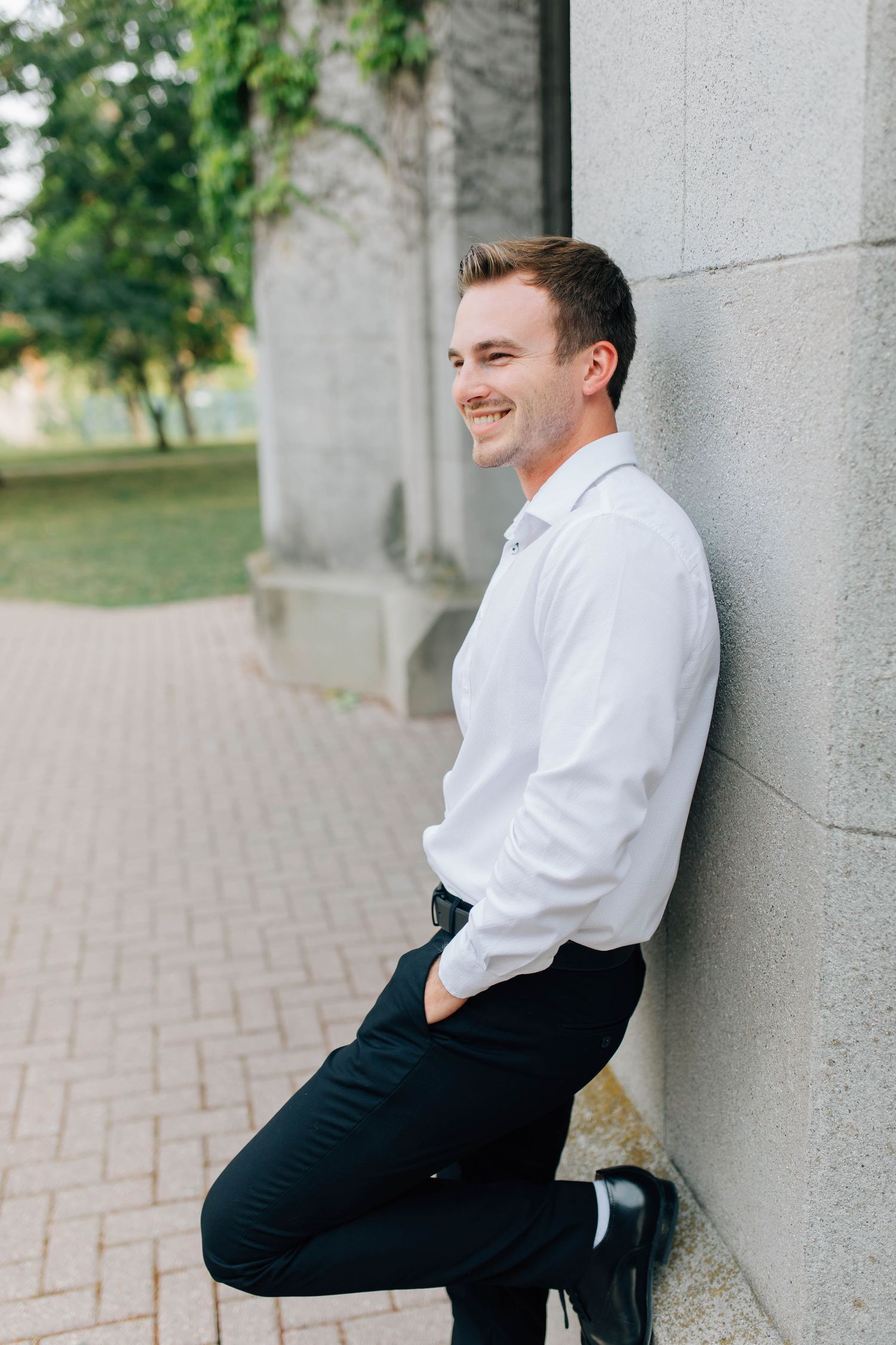 Handsome smiling groom in white shirt leaning against a stone column in an outdoor garden setting.