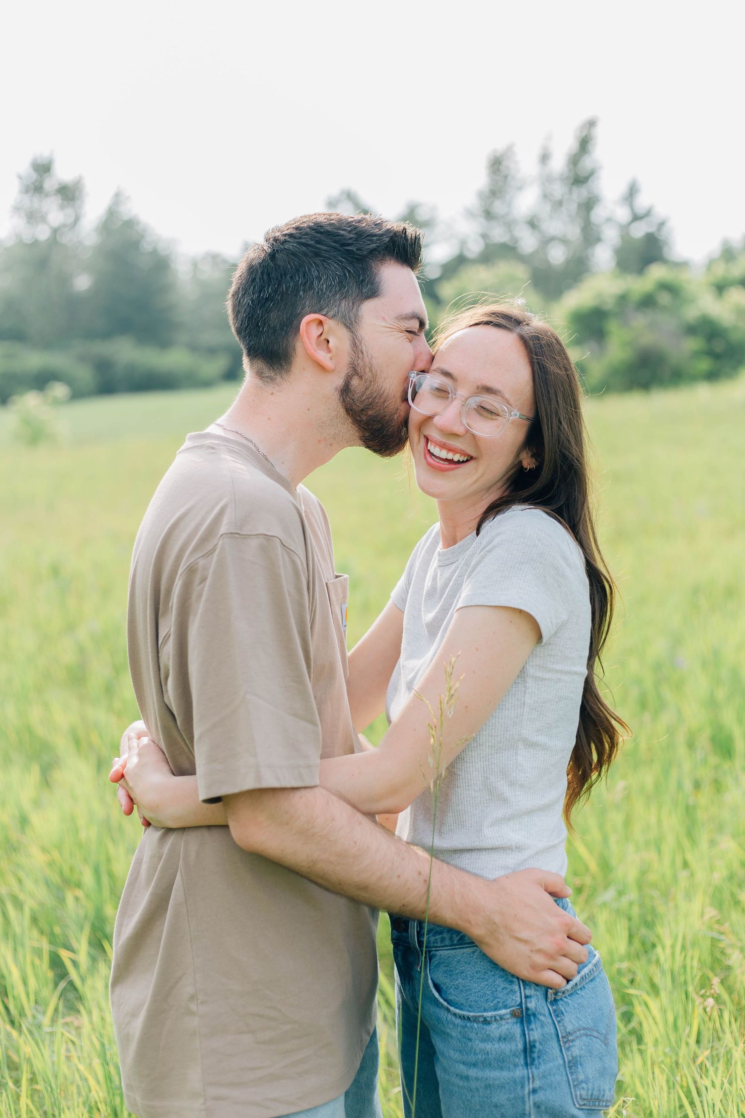 Man kissing laughing womans cheek while embracing in sunny green meadow outdoors.