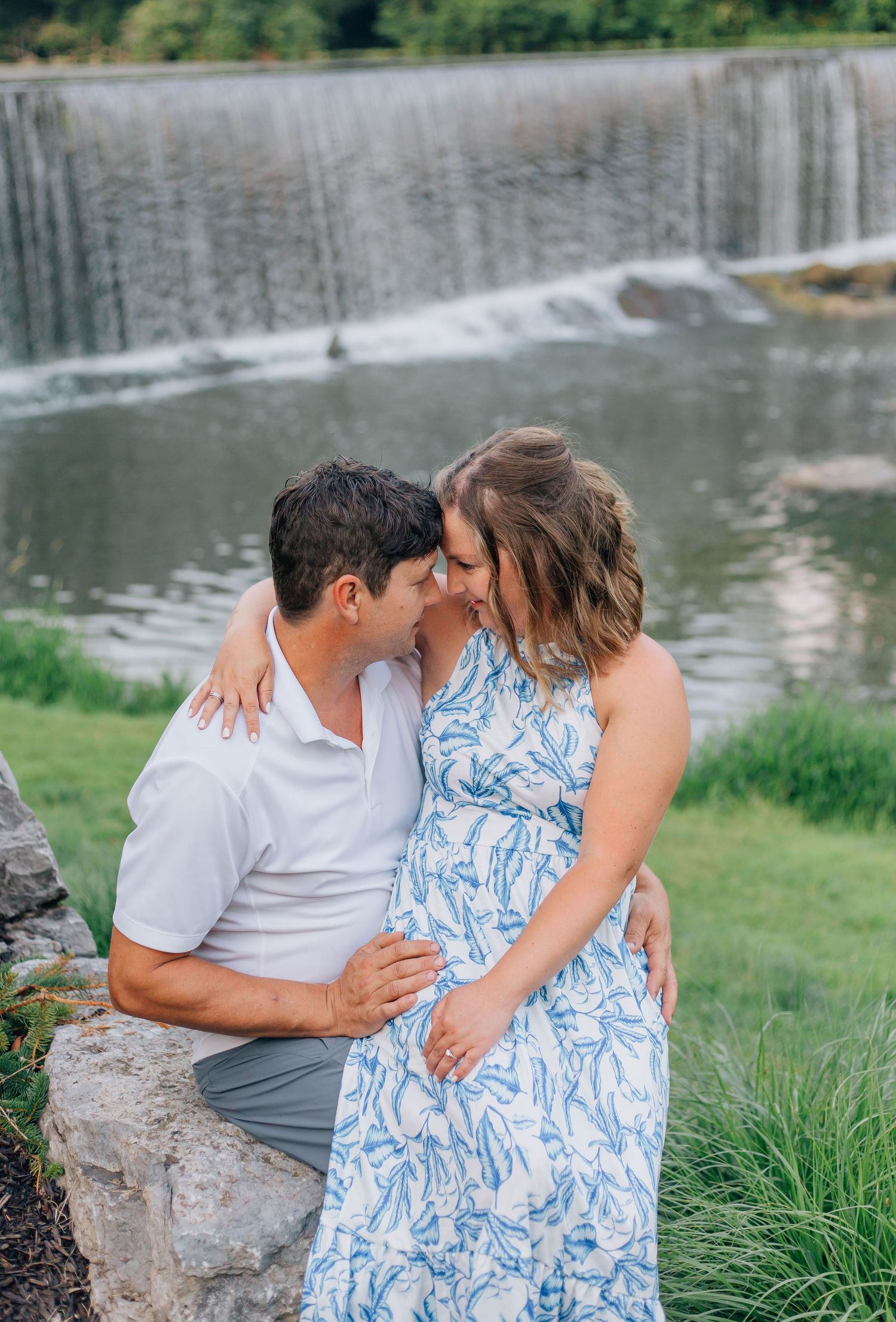 Couple sharing a tender moment by a scenic waterfall, woman in blue floral dress embracing man in white polo.