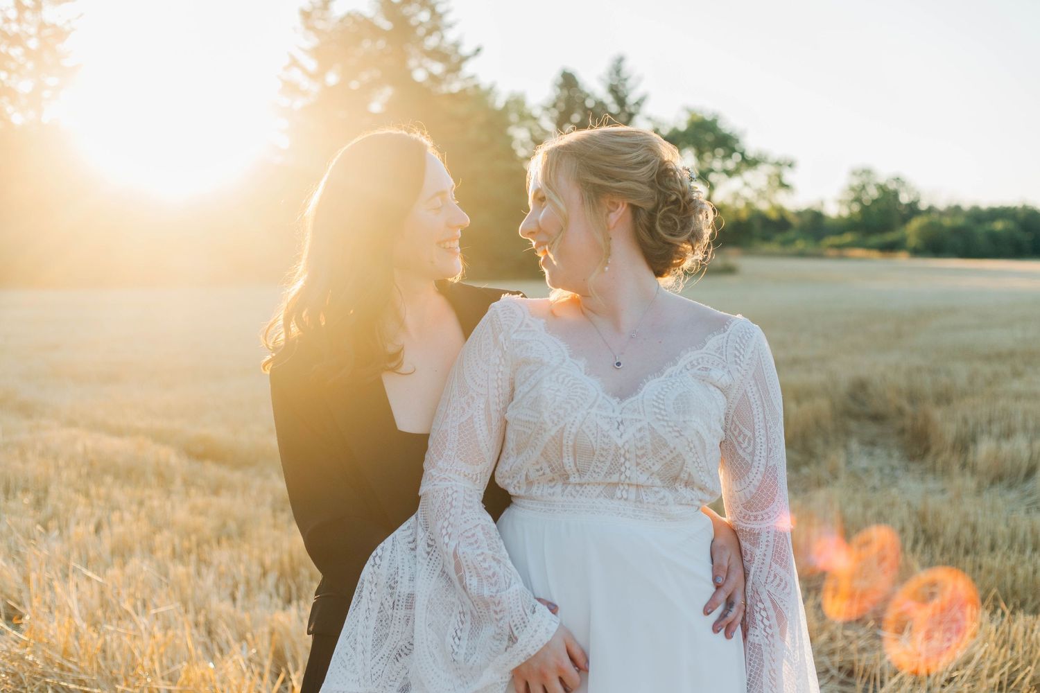 Lesbian wedding couple smiling at each other in a sun-drenched golden wheat field.