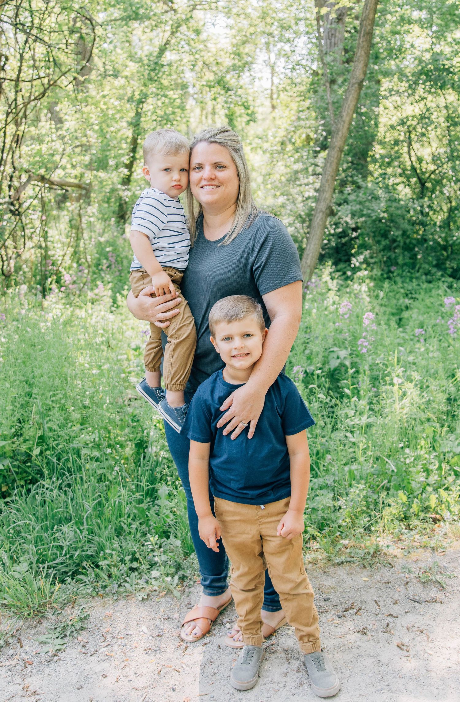 Smiling mother holding a toddler while standing with her young son outdoors in nature.