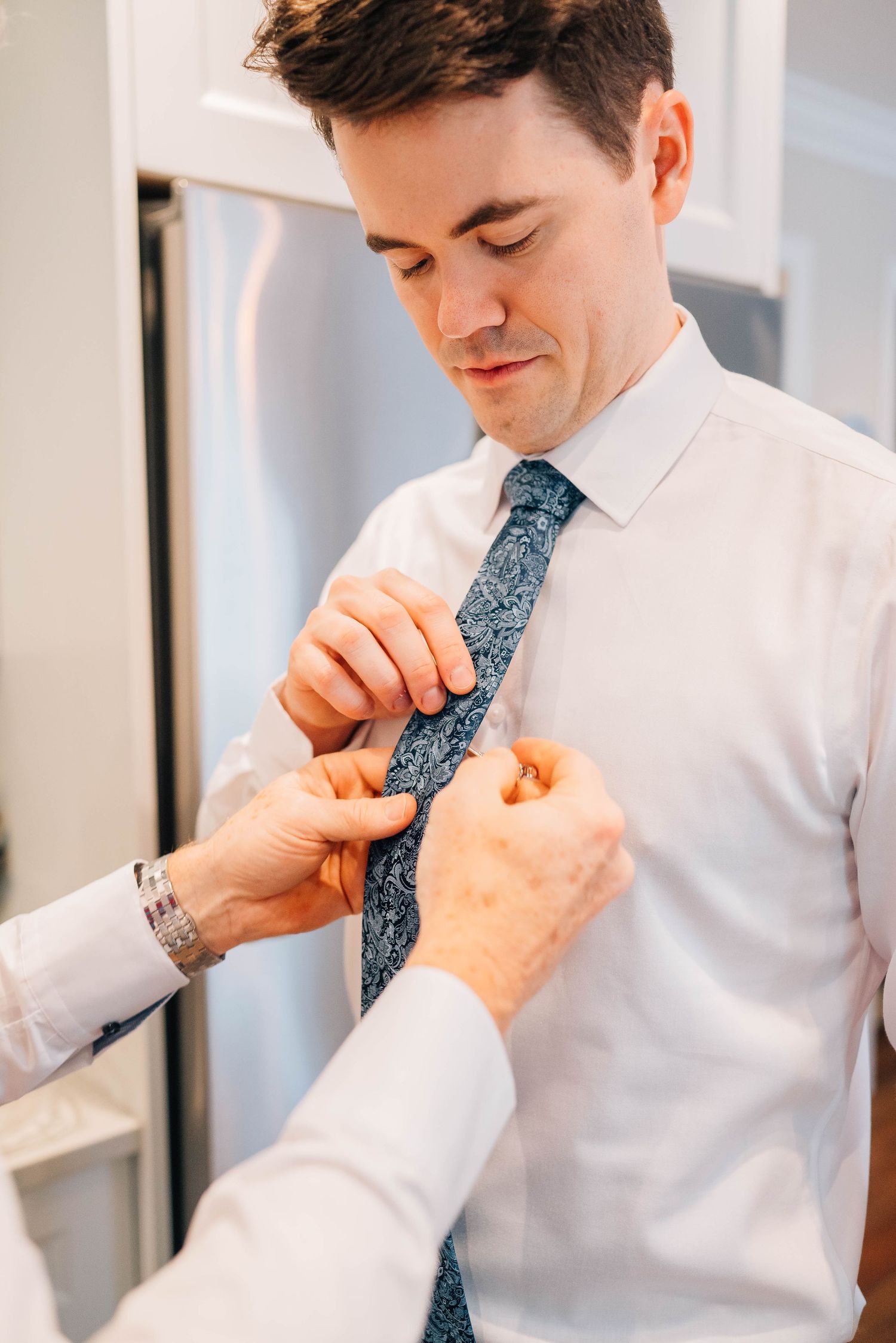 Groom having his blue paisley tie adjusted by another person while preparing for his wedding.