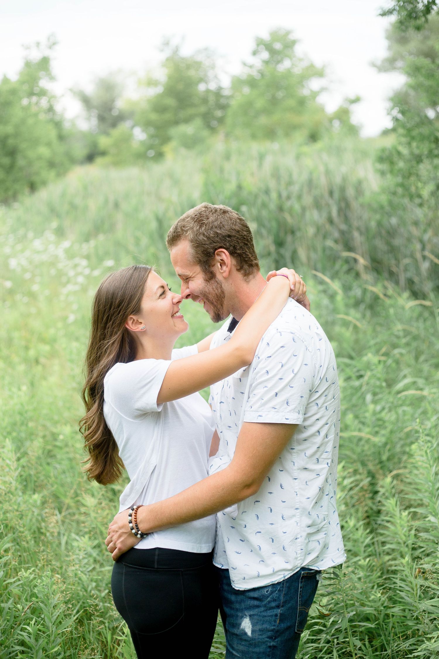 A joyful couple embraces and smiles at each other surrounded by lush green meadow grass.