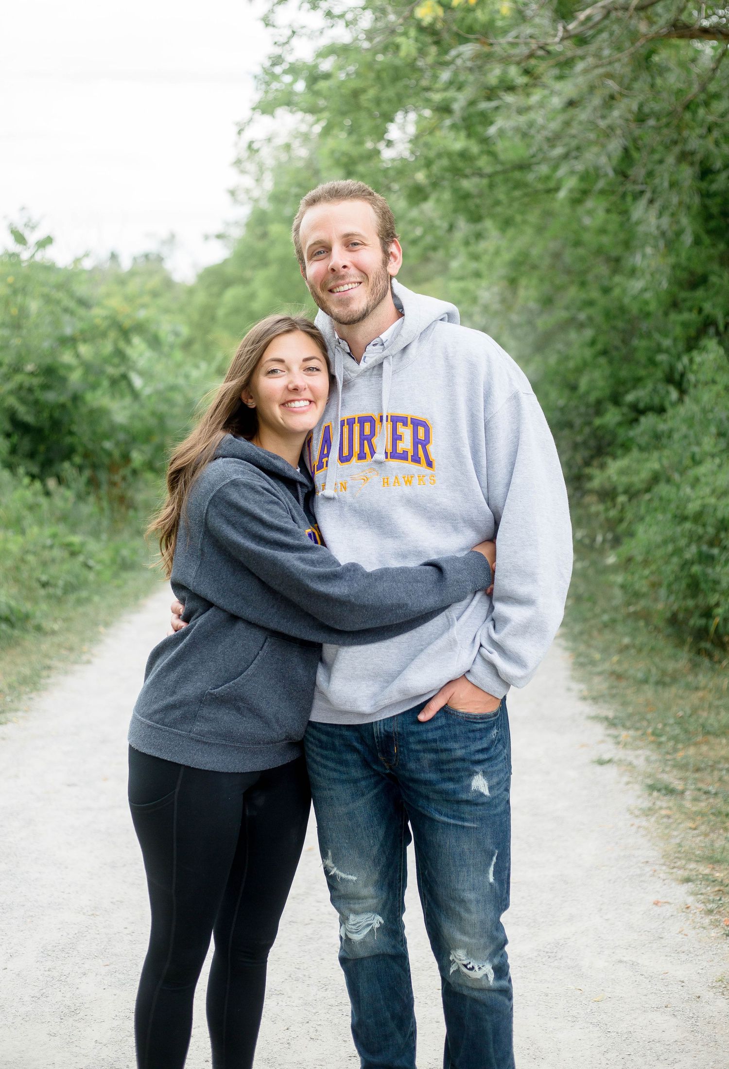 Couple embracing and smiling while wearing Laurier University hoodies on a scenic outdoor gravel trail.