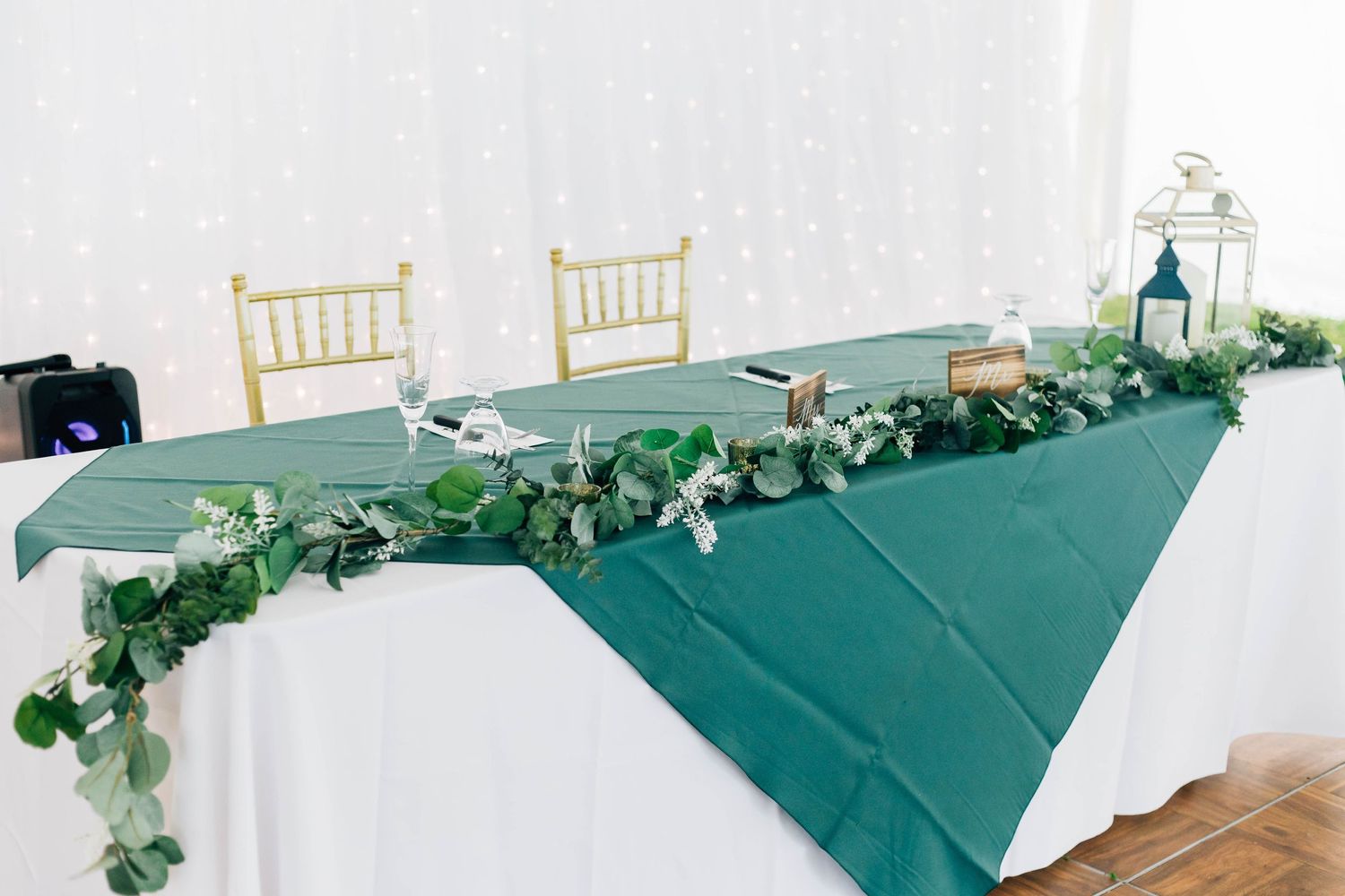 Elegant wedding head table with green tablecloth, eucalyptus garland, and gold chiavari chairs.