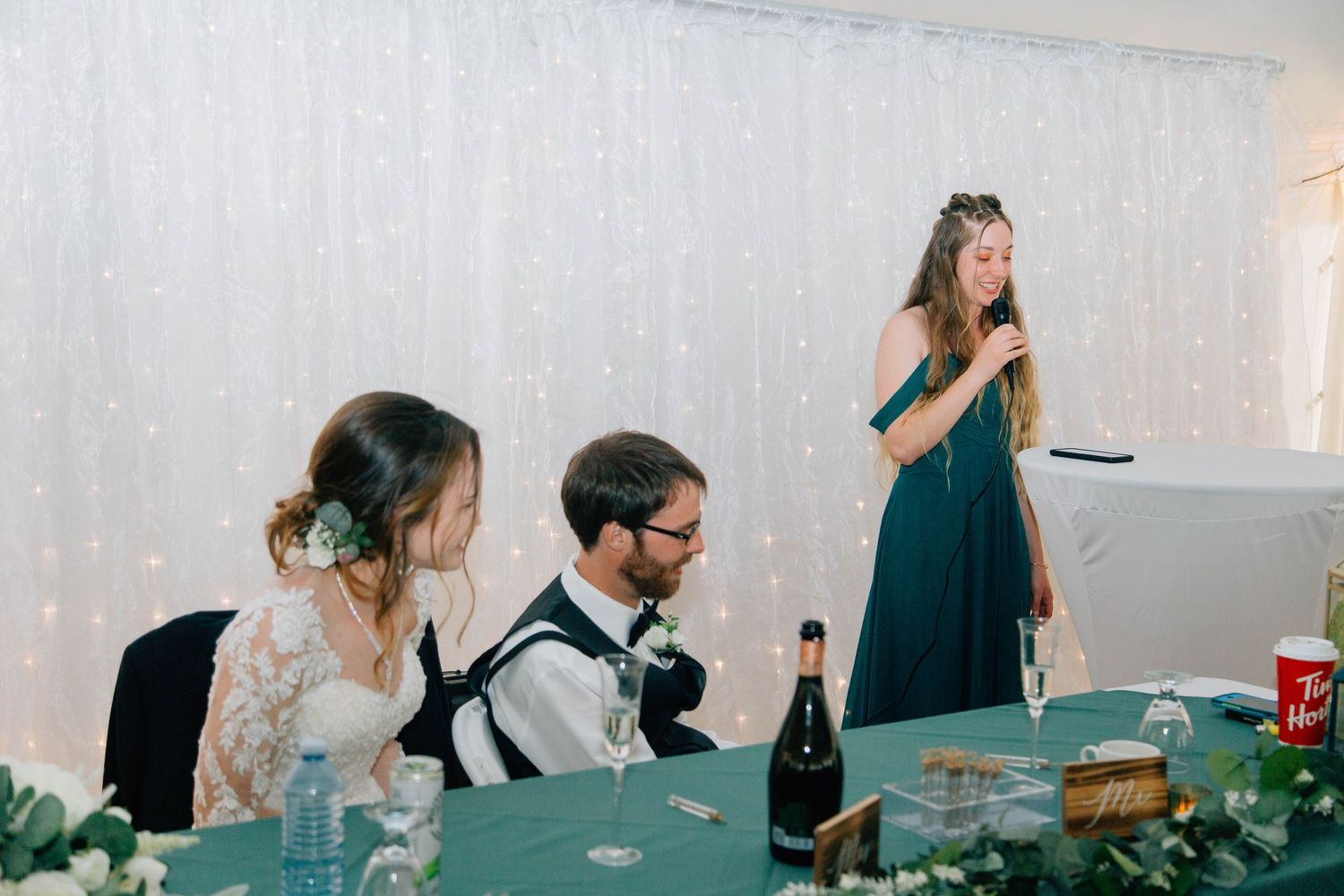 Bridesmaid in teal dress gives wedding toast speech while bride and groom sit at decorated reception table.