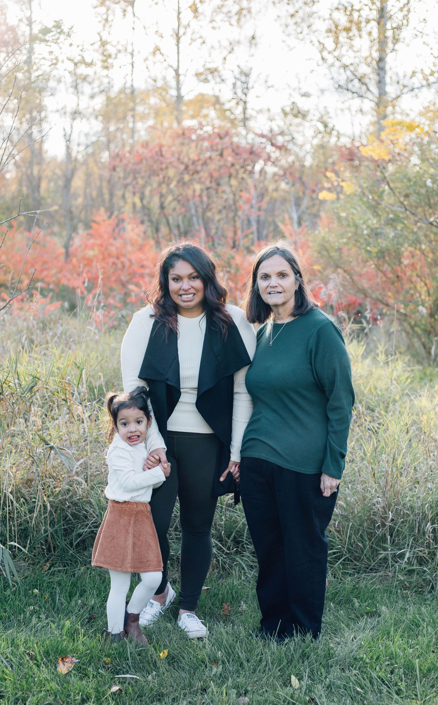 Woman and older lady smiling with young girl in rust skirt among vibrant fall foliage.