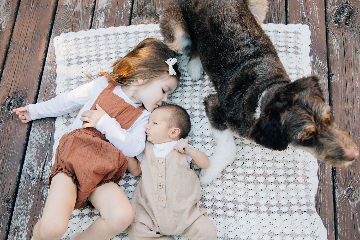 Little girl and newborn baby lying on blanket with large fluffy dog on wooden deck.