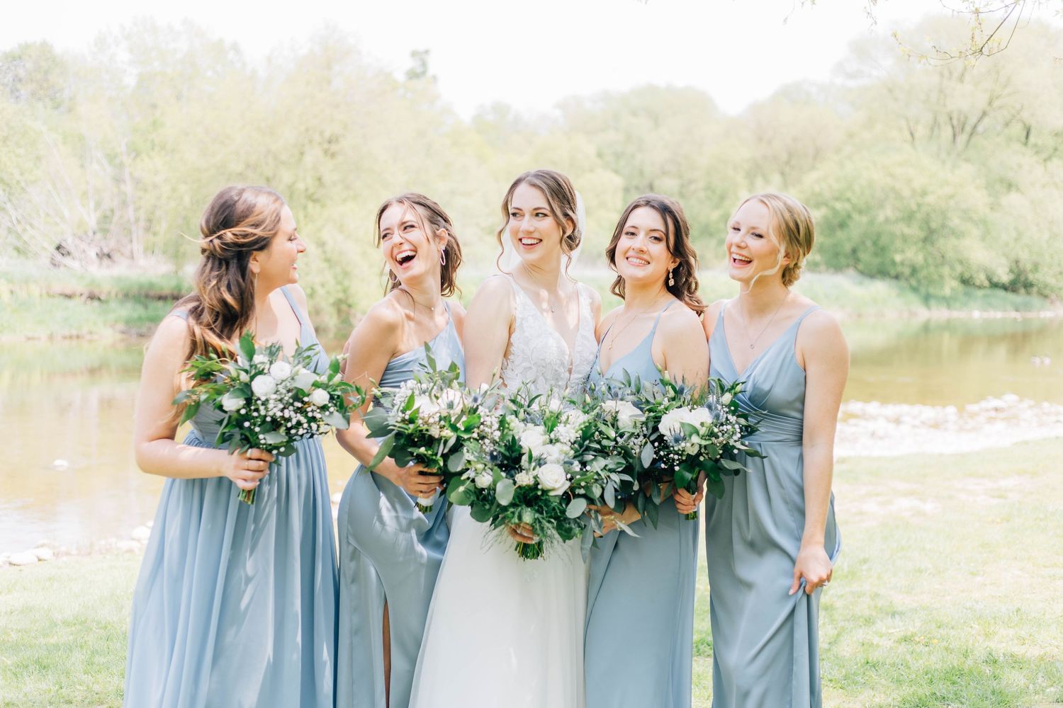 Bride and bridesmaids in light blue dresses laugh joyfully while holding lush white and green bouquets.