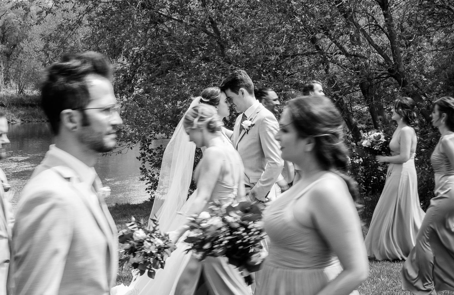 Black and white candid photo of bride in veil laughing with wedding party walking through lush greenery.