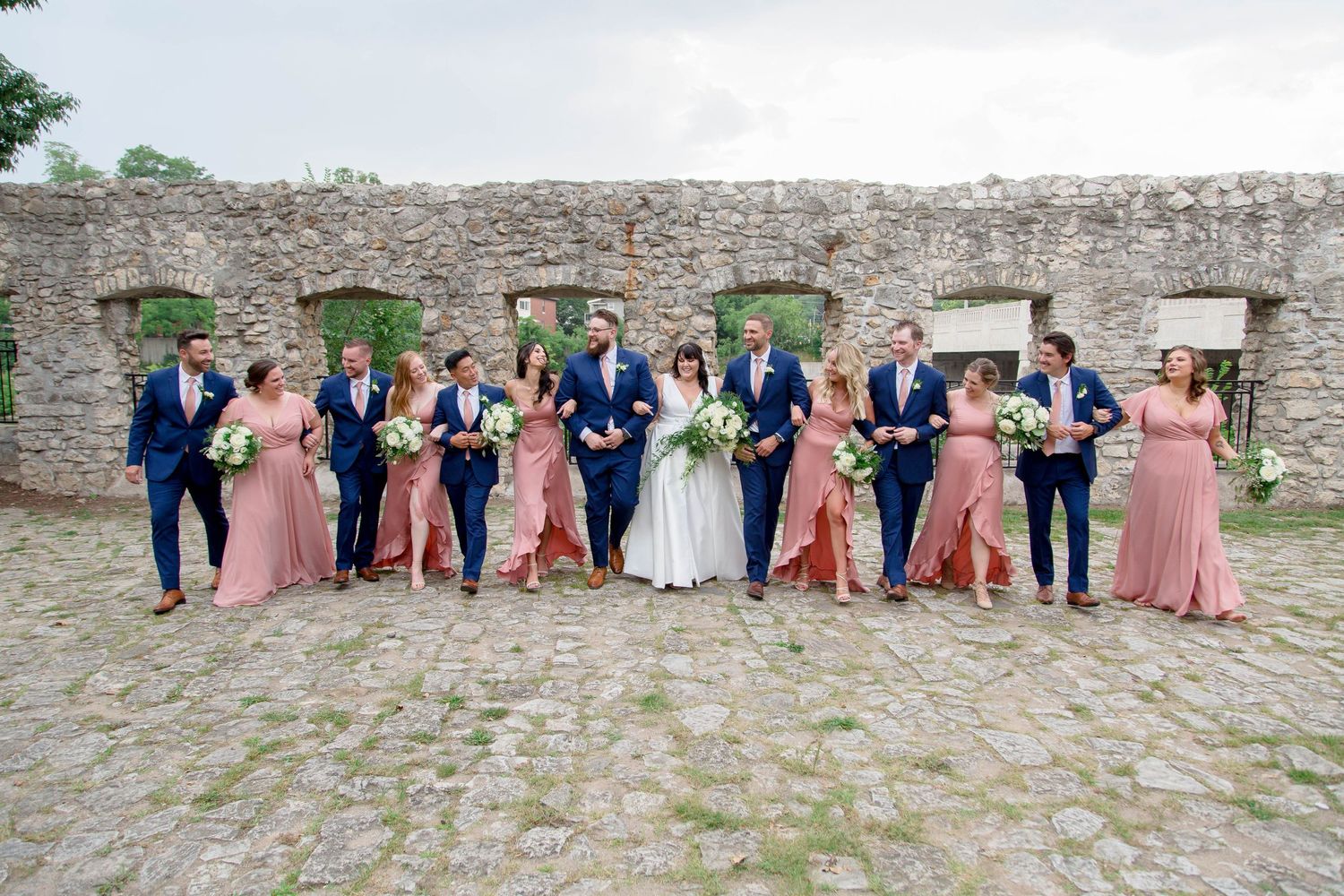 A wedding party in navy suits and dusty rose gowns walks together in front of a stone wall.