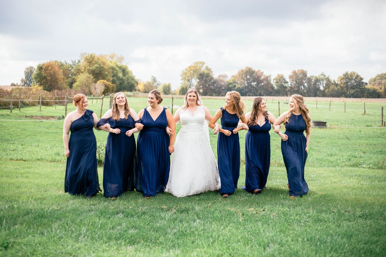 Bride and six bridesmaids in navy dresses walk arm-in-arm across a green field on wedding day.