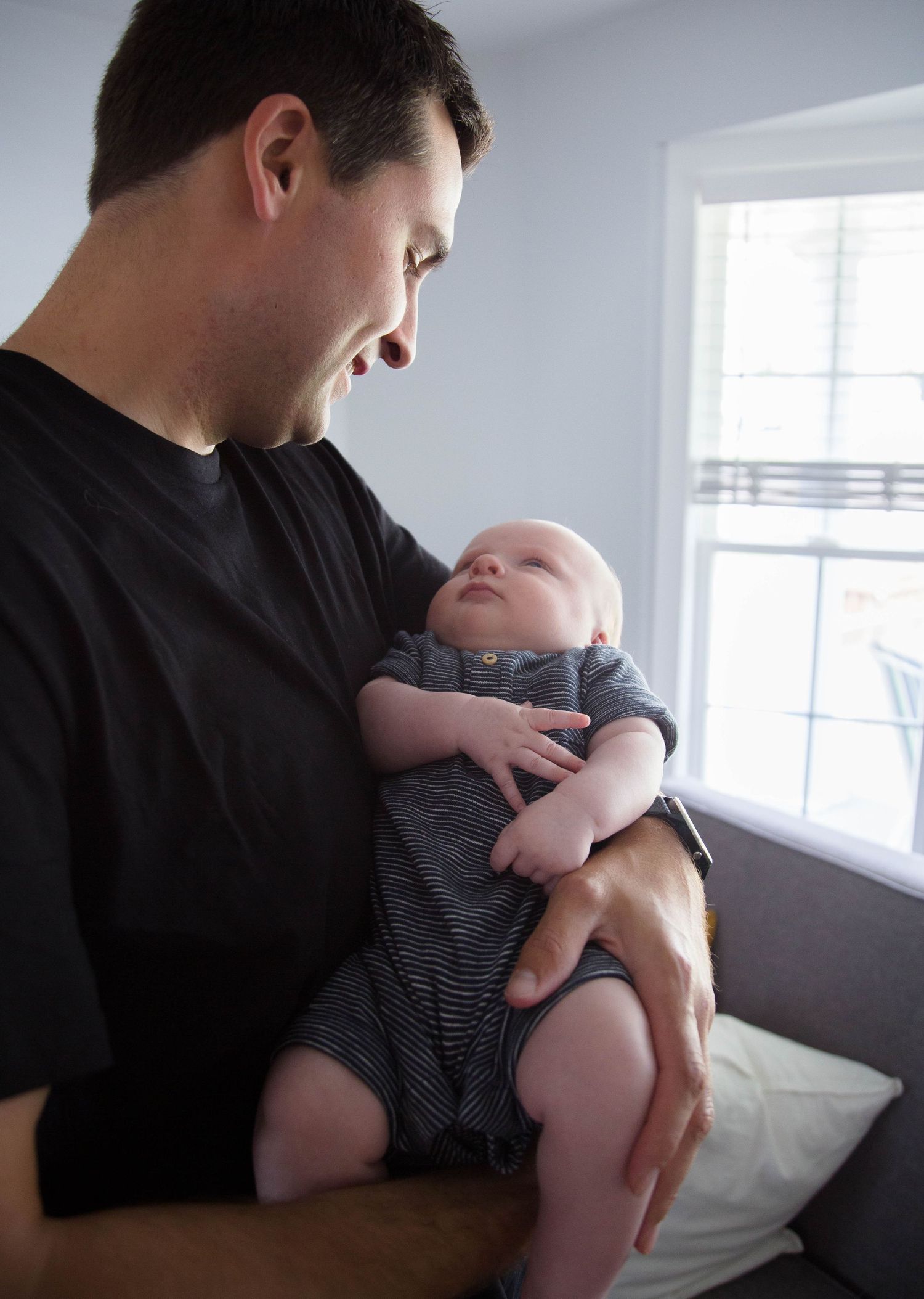 Father in black shirt gazing lovingly at newborn baby near bright window at home.