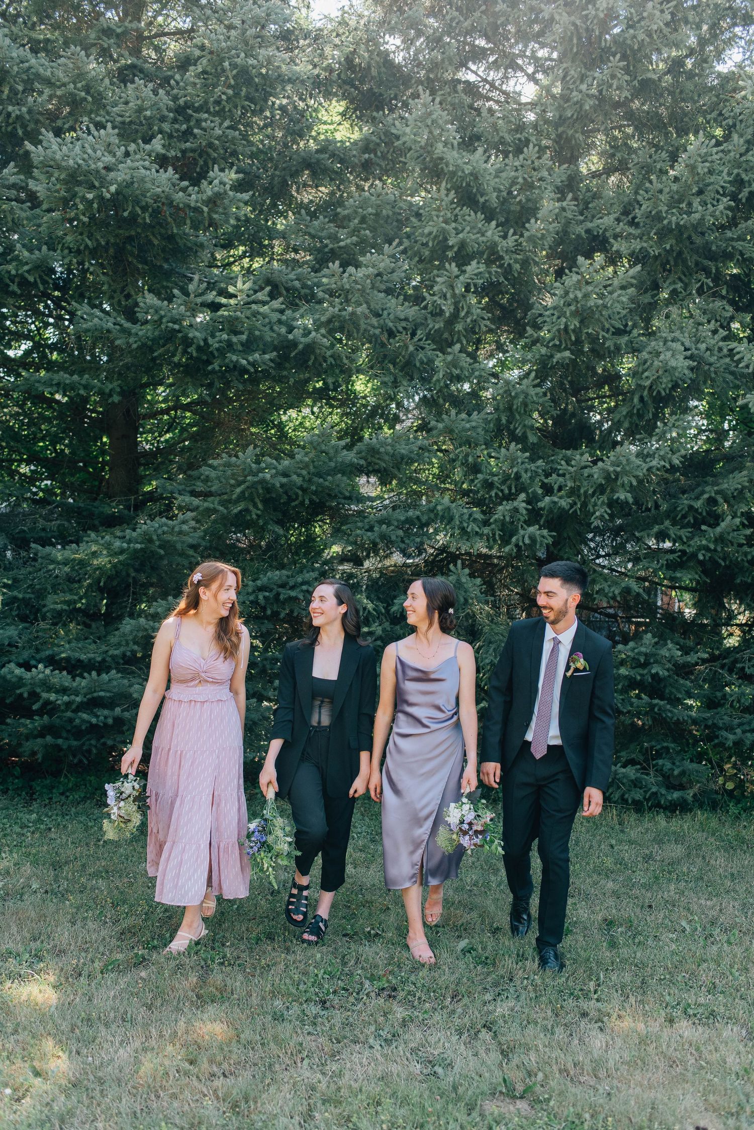 Wedding party of four walks across grass before tall evergreen trees holding wildflower bouquets.
