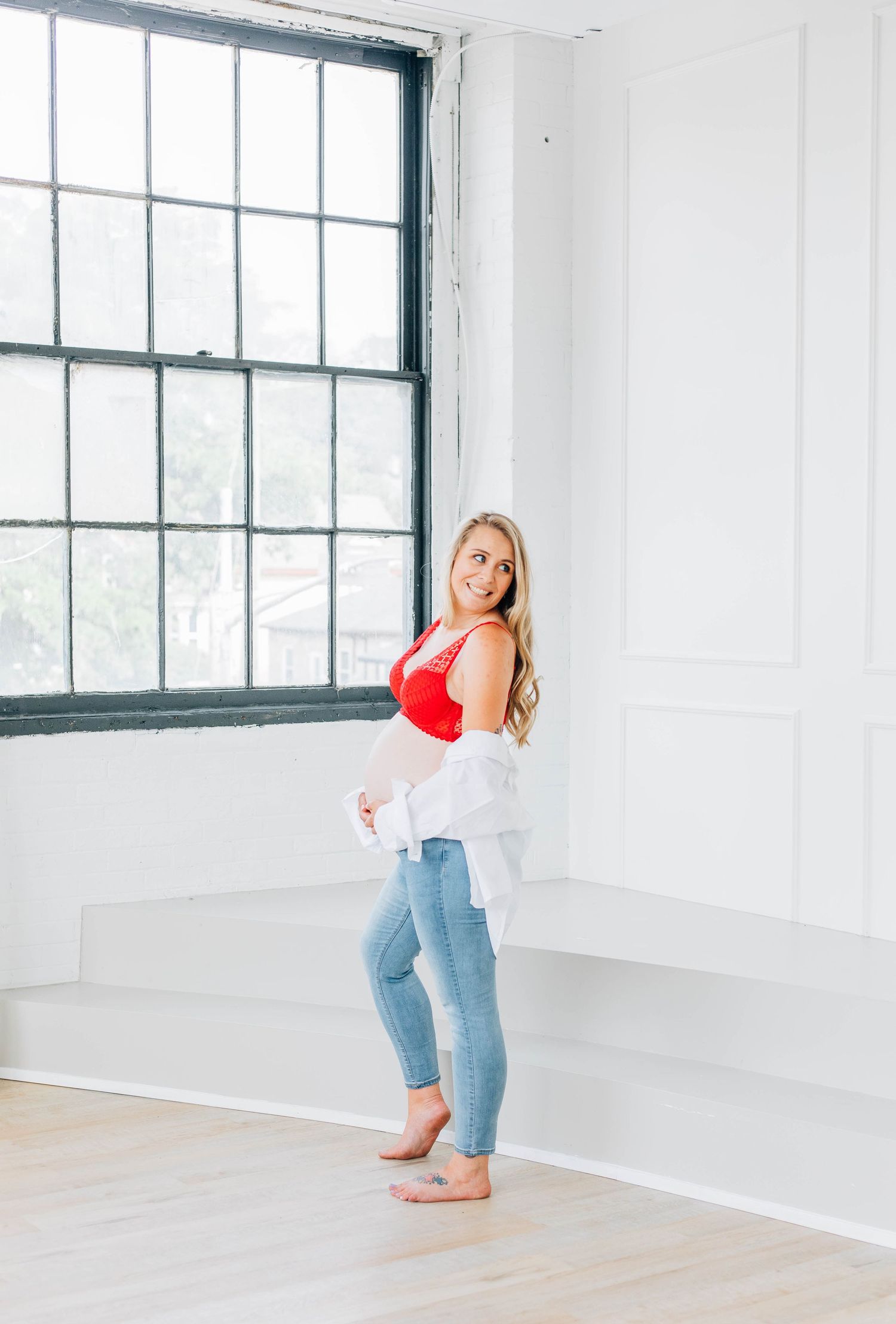 Side profile of pregnant blonde woman in red bralette and jeans standing by large studio window.