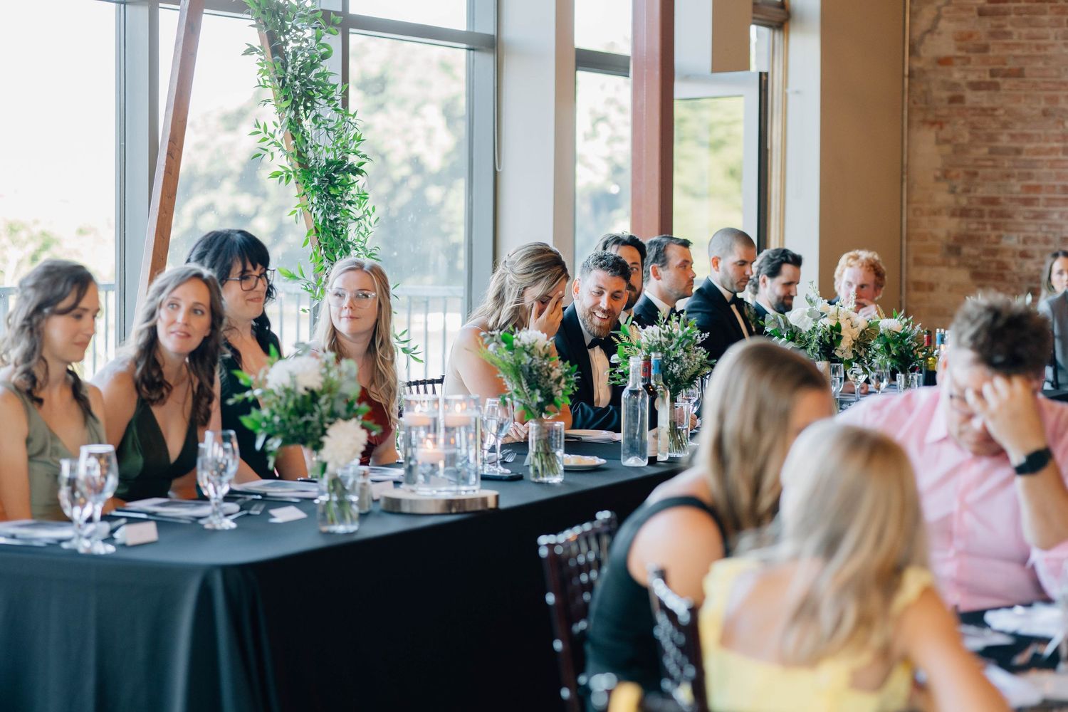 Wedding reception guests seated at long black tables near large windows in a bright industrial venue.