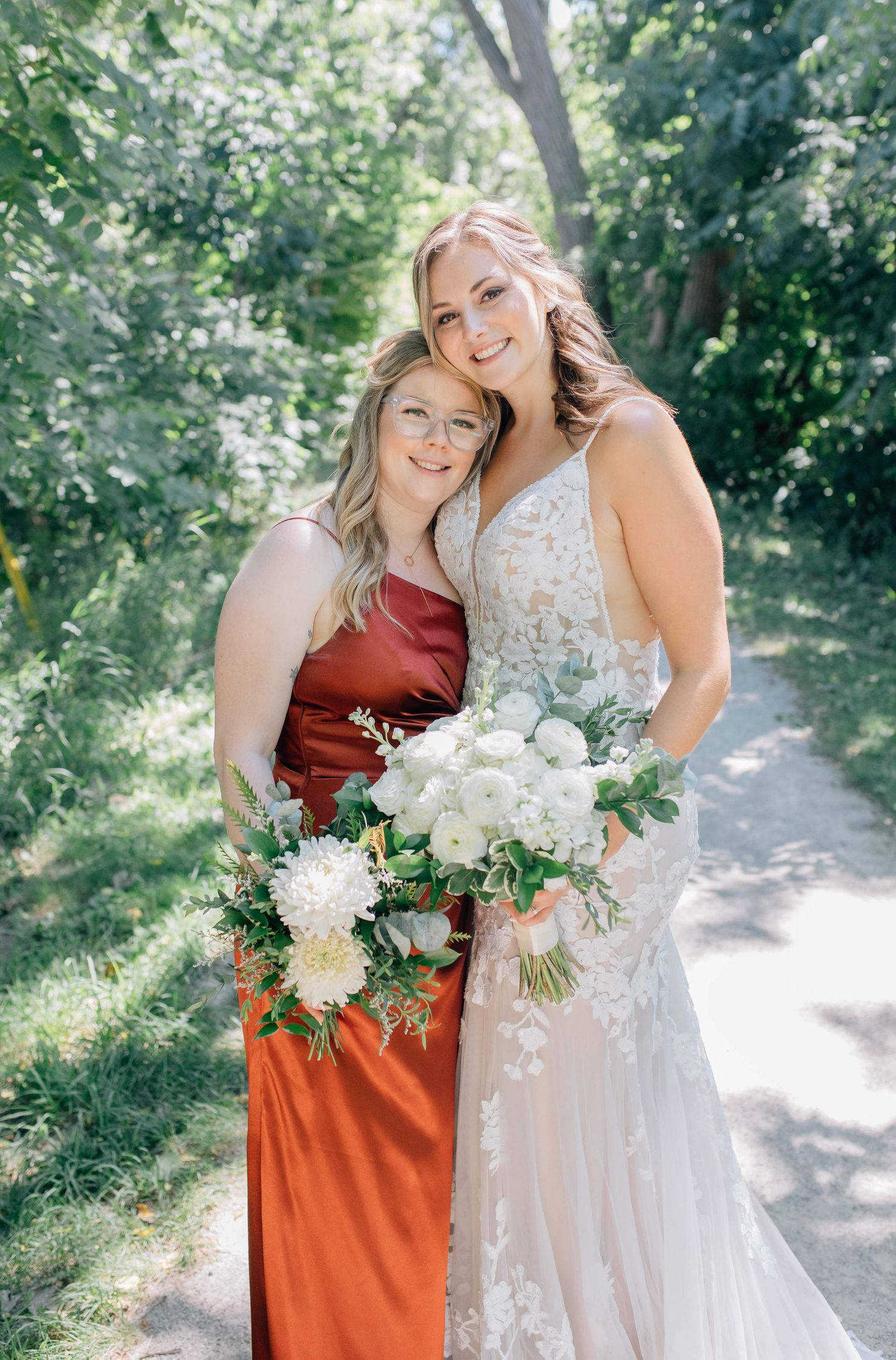 Bride in lace wedding gown embraces bridesmaid in rust-red dress while holding white floral bouquets outdoors.