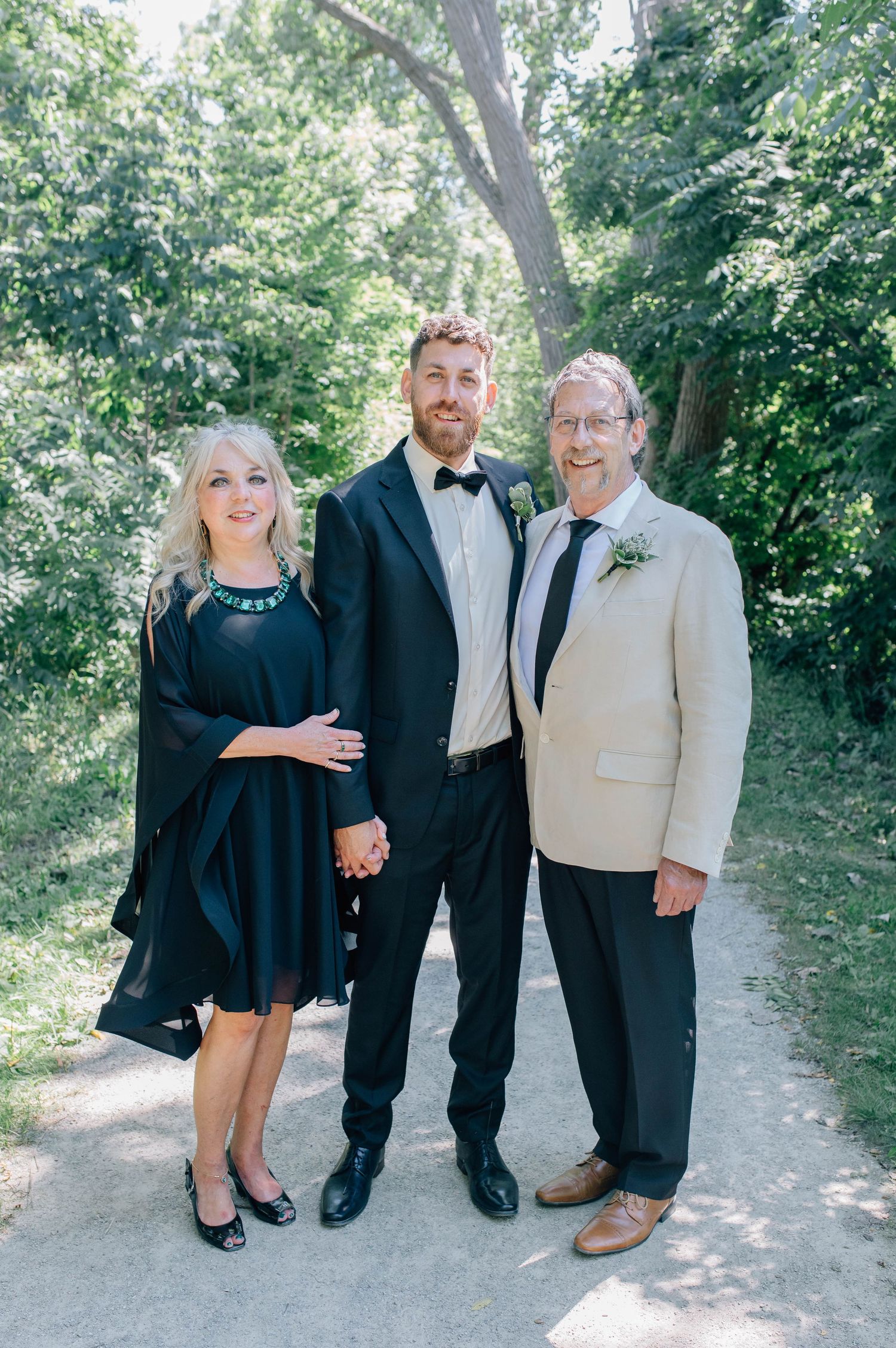 Groom in a black tuxedo posing with his parents on a sunny tree-lined path on his wedding day.