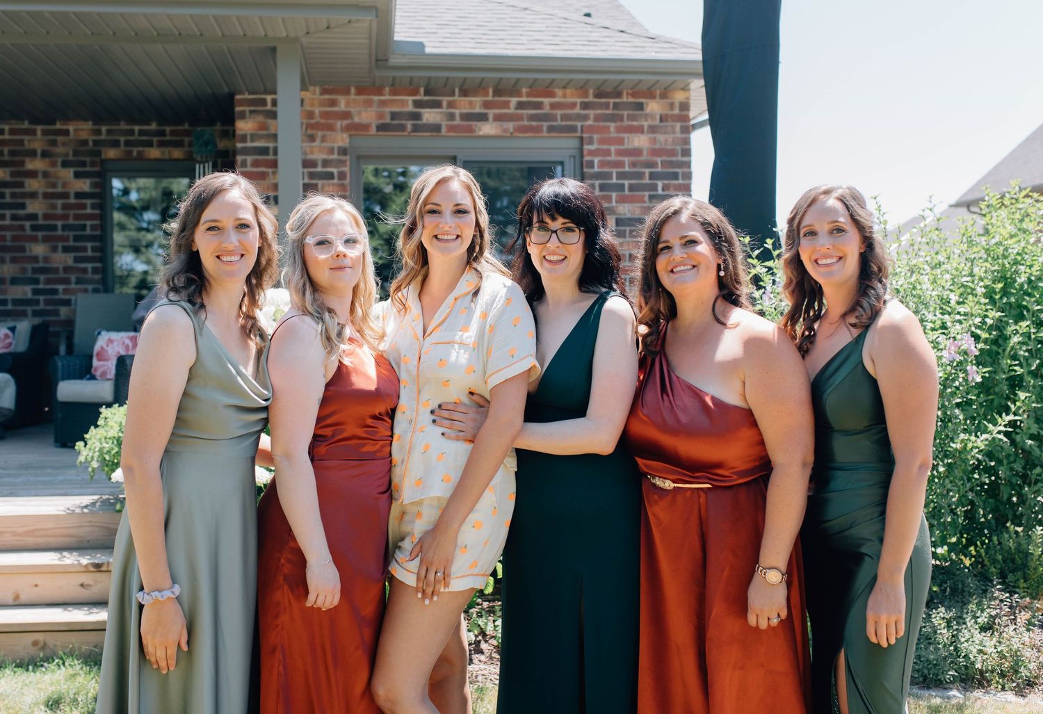 Bride in white robe posing outdoors with six bridesmaids in earth-toned dresses.