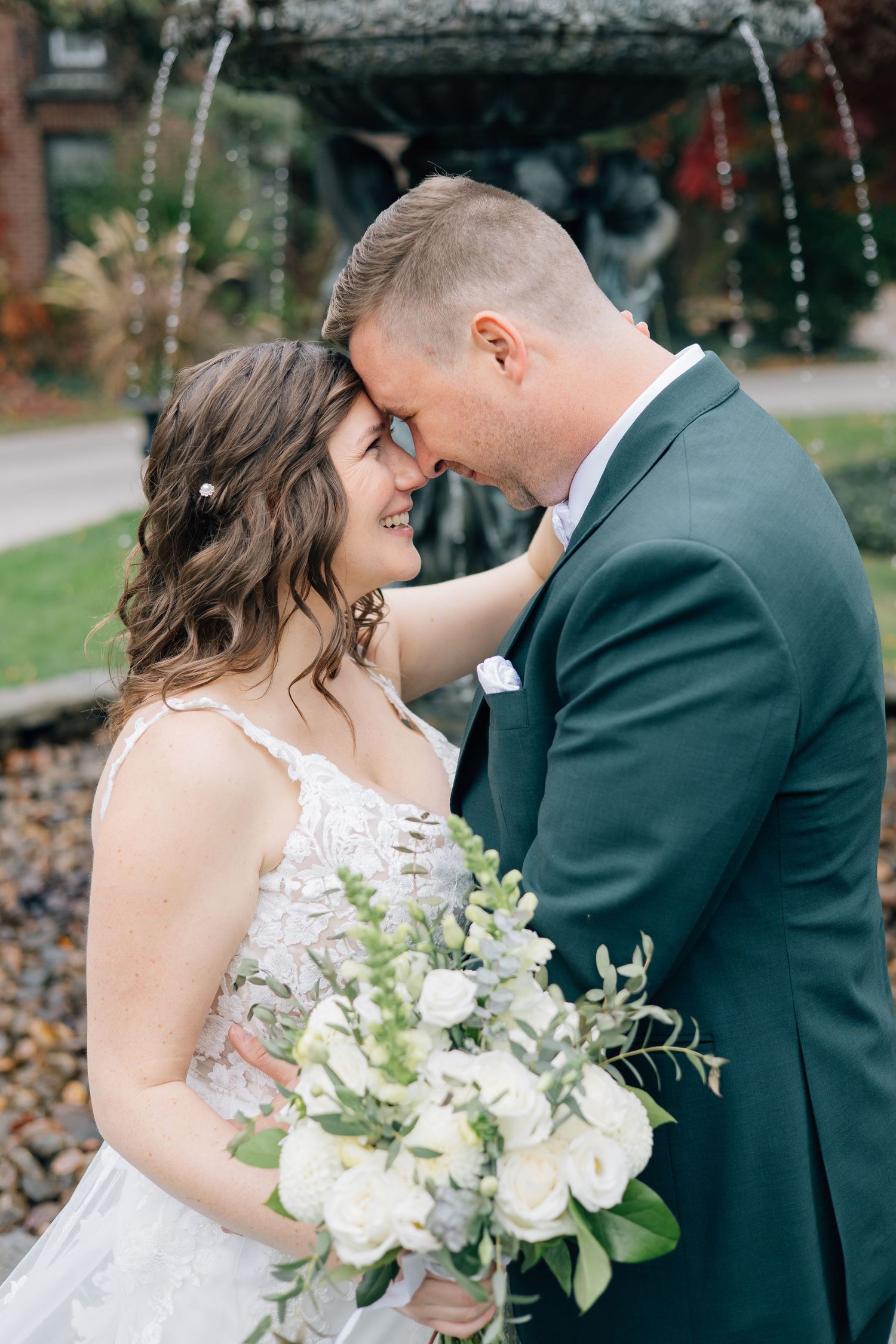 Bride and groom touching foreheads near fountain, bride holding white floral bouquet.