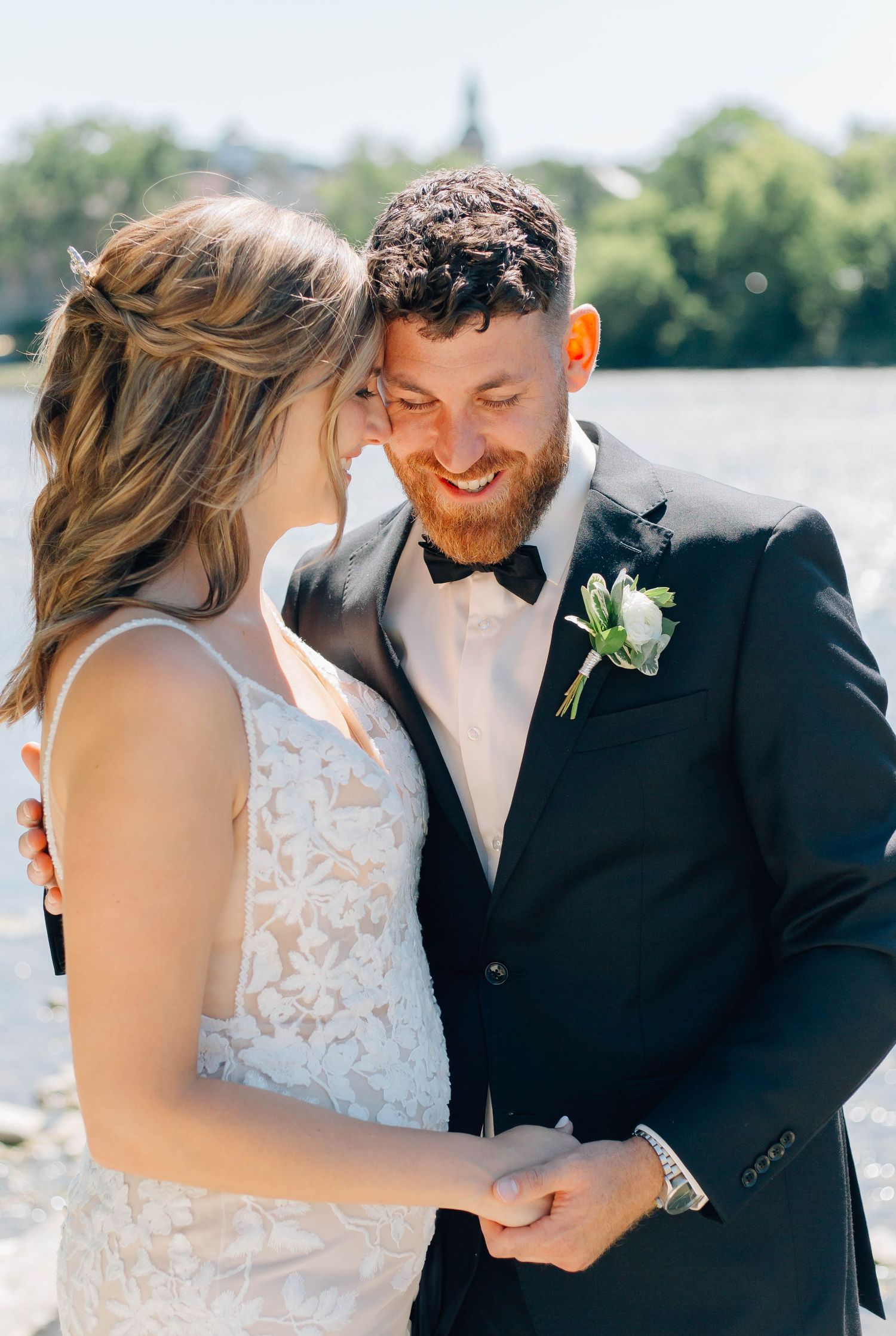 Bride and groom sharing a laugh by the water, smiling and touching foreheads in wedding attire.