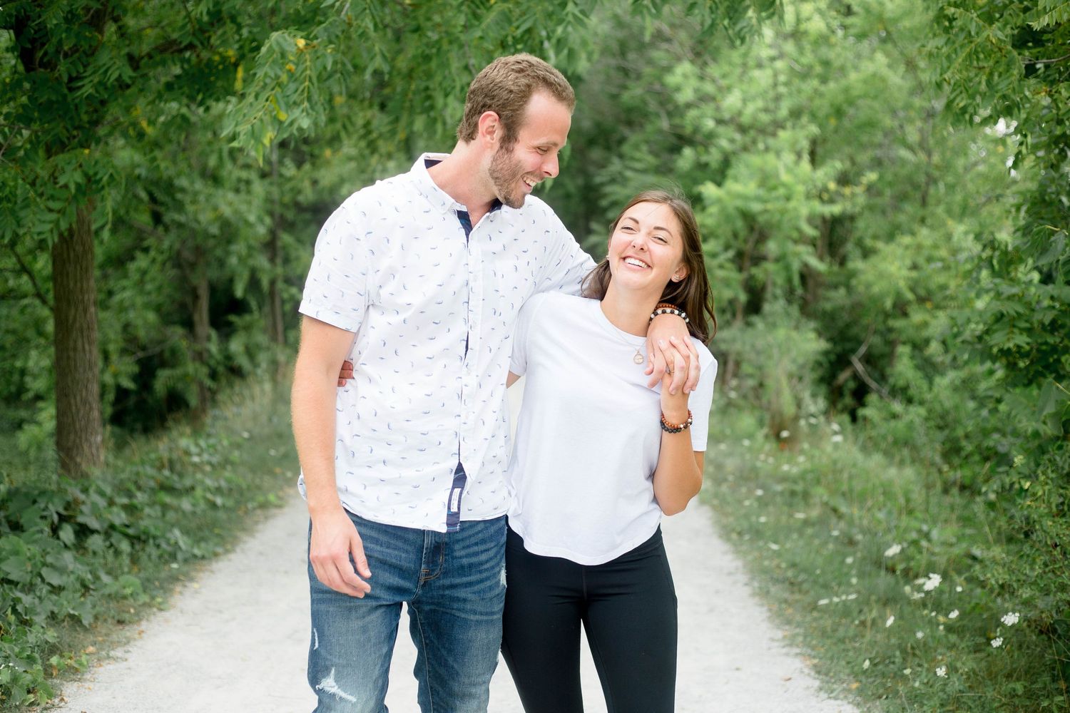 A smiling couple walks closely together along a tree-lined path during an outdoor photo session.