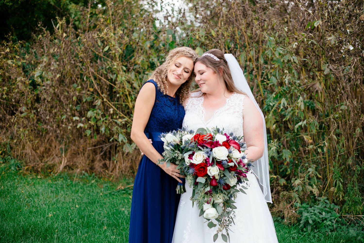 Bride and bridesmaid in navy lace dress share a tender moment holding a lush red and white bouquet.