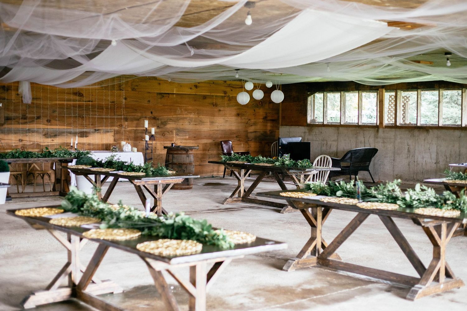 Rustic barn wedding reception venue with wooden tables decorated with greenery garlands and draped fabric ceiling.