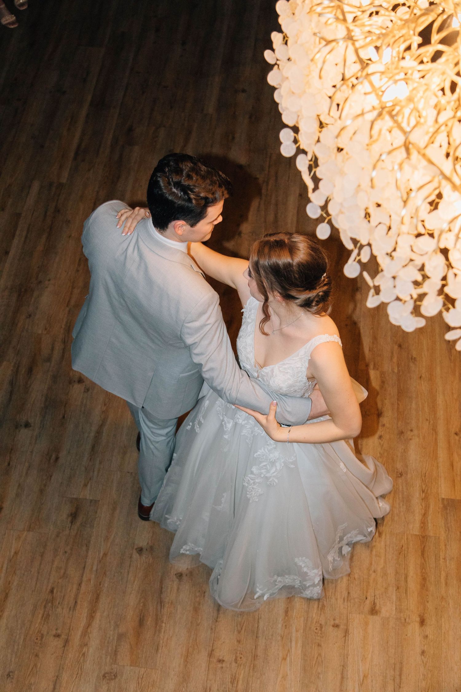 Aerial view of bride in lace gown and groom dancing beneath elegant chandelier at reception.