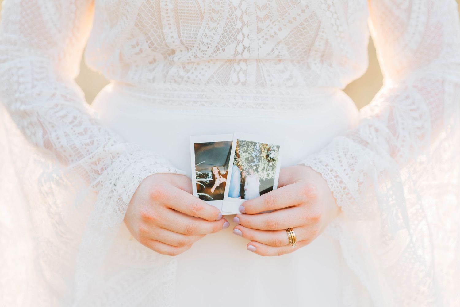 Close-up of bride in white lace dress holding two Polaroid photos with delicate ring on finger.