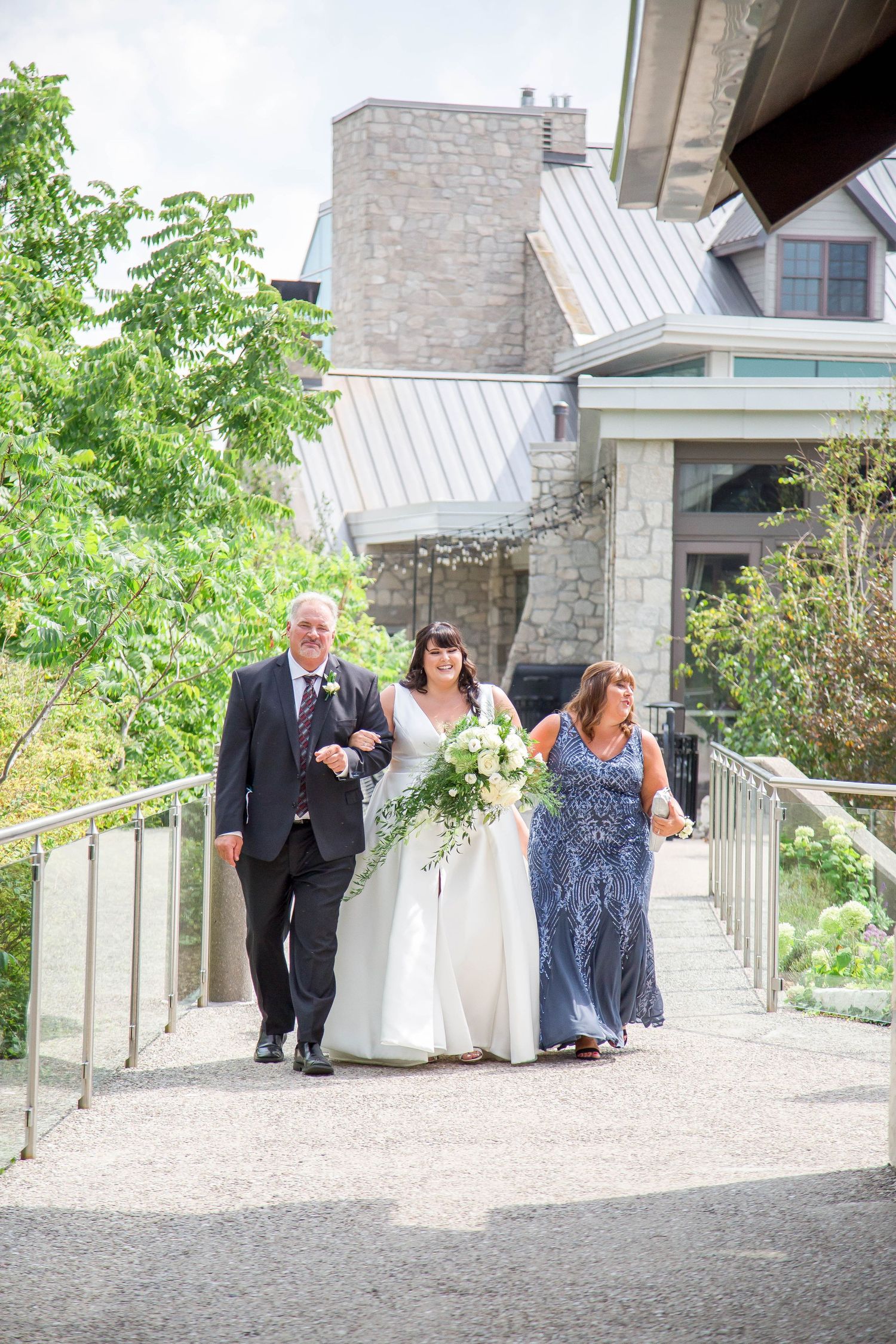 A bride in a white gown with a cascading bouquet walks with her parents toward a stone venue.