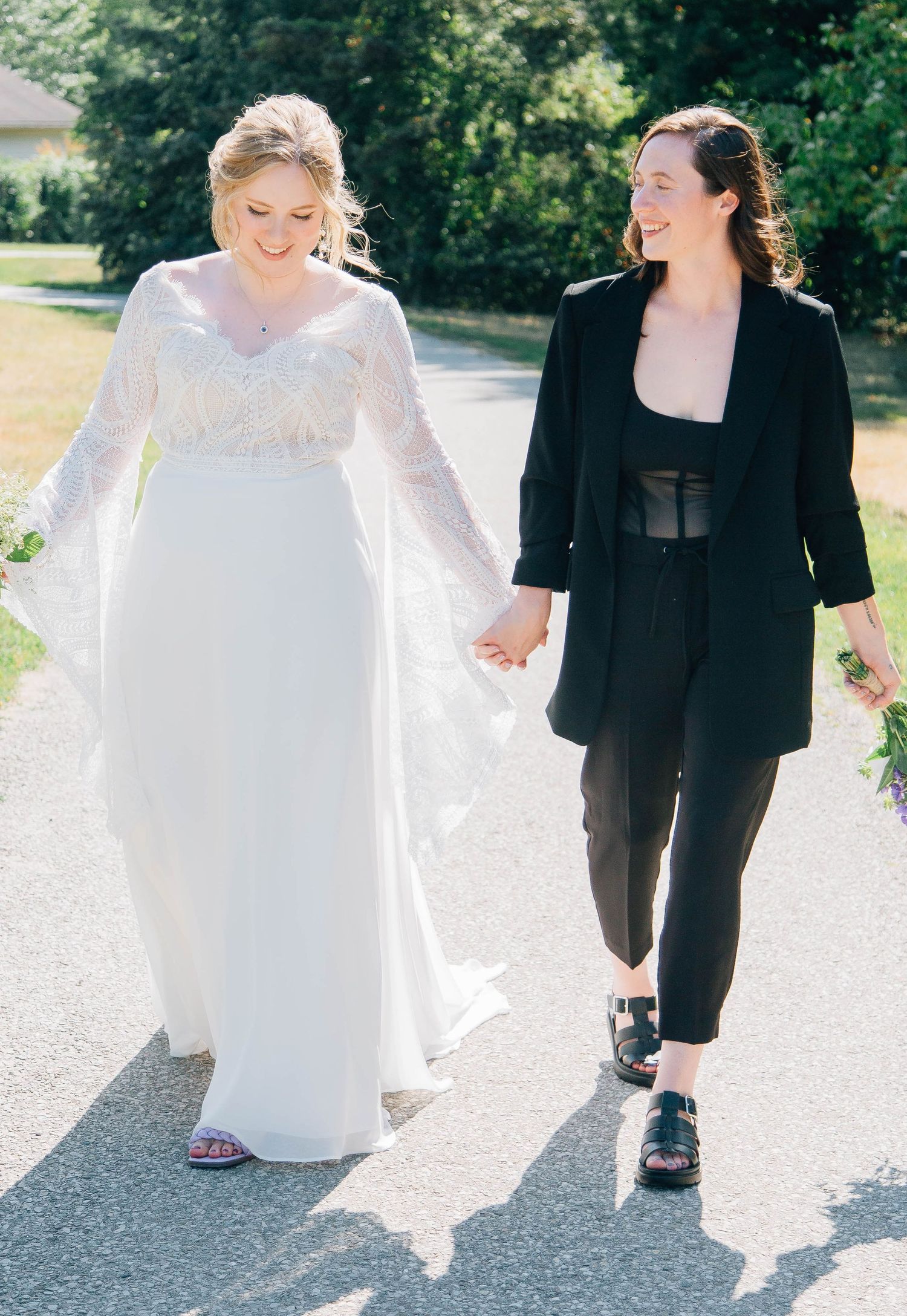 Bride in lace gown and woman in black blazer walk hand in hand along a sunny outdoor path.