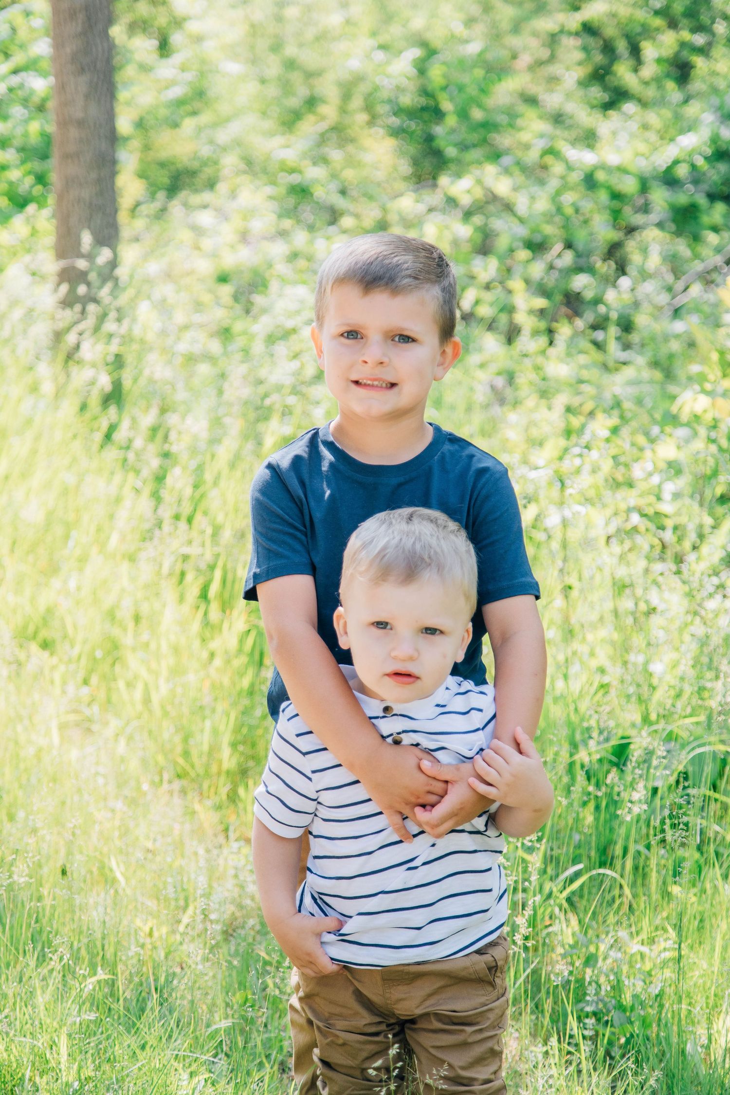 Older boy hugging younger toddler brother while standing in a bright sunny green field.