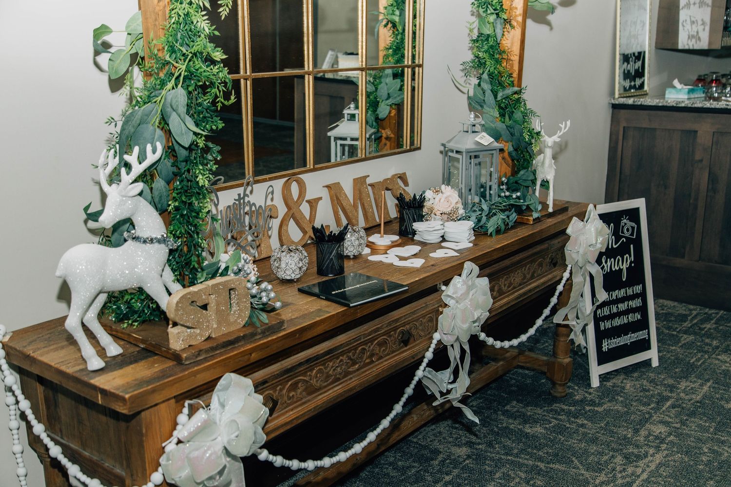 White glitter reindeer figurine beside gold SD letters and silver berry garland on wooden wedding display.