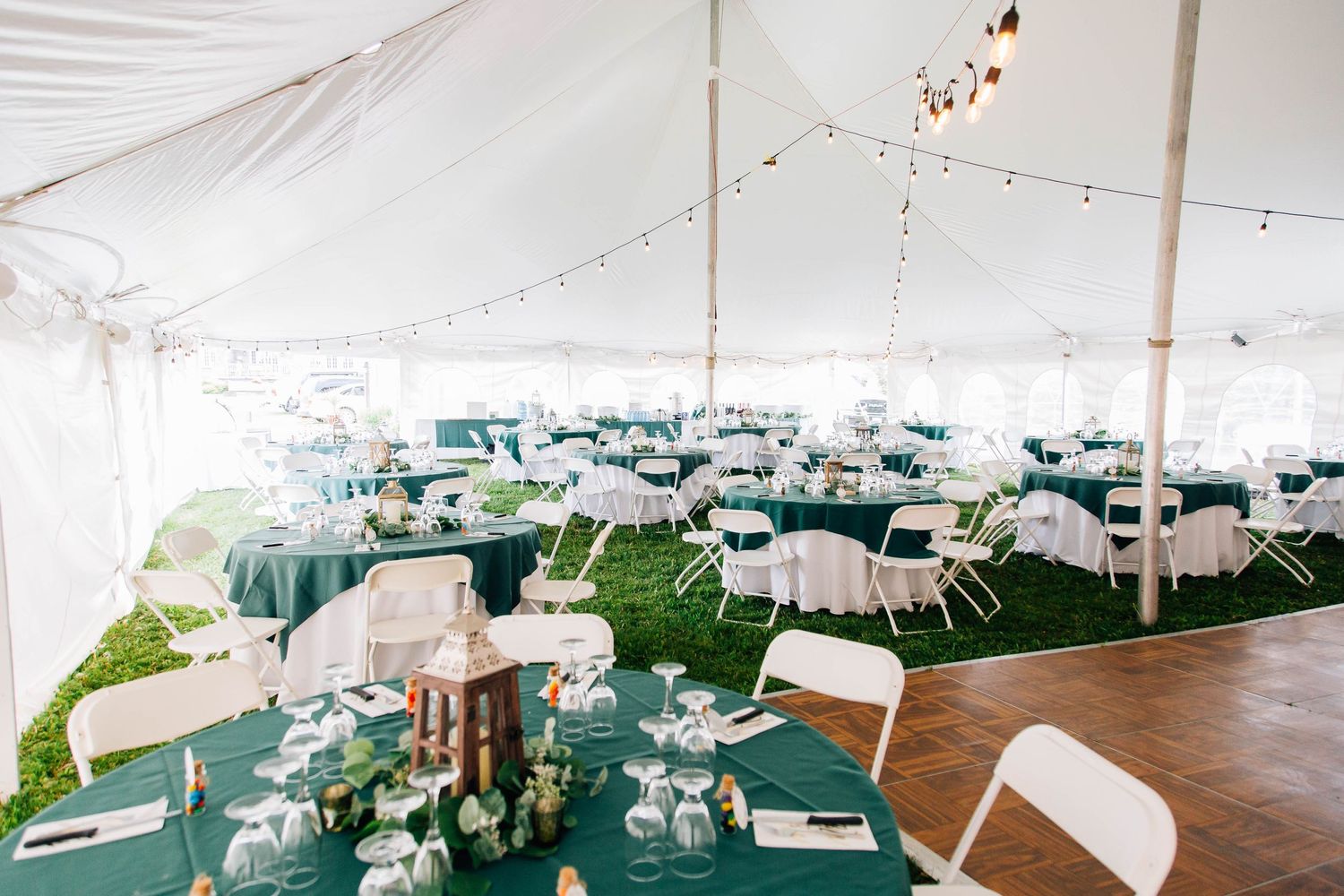 Elegant wedding reception tent with green tablecloths, white chairs, string lights, and floral centerpieces on lush grass.