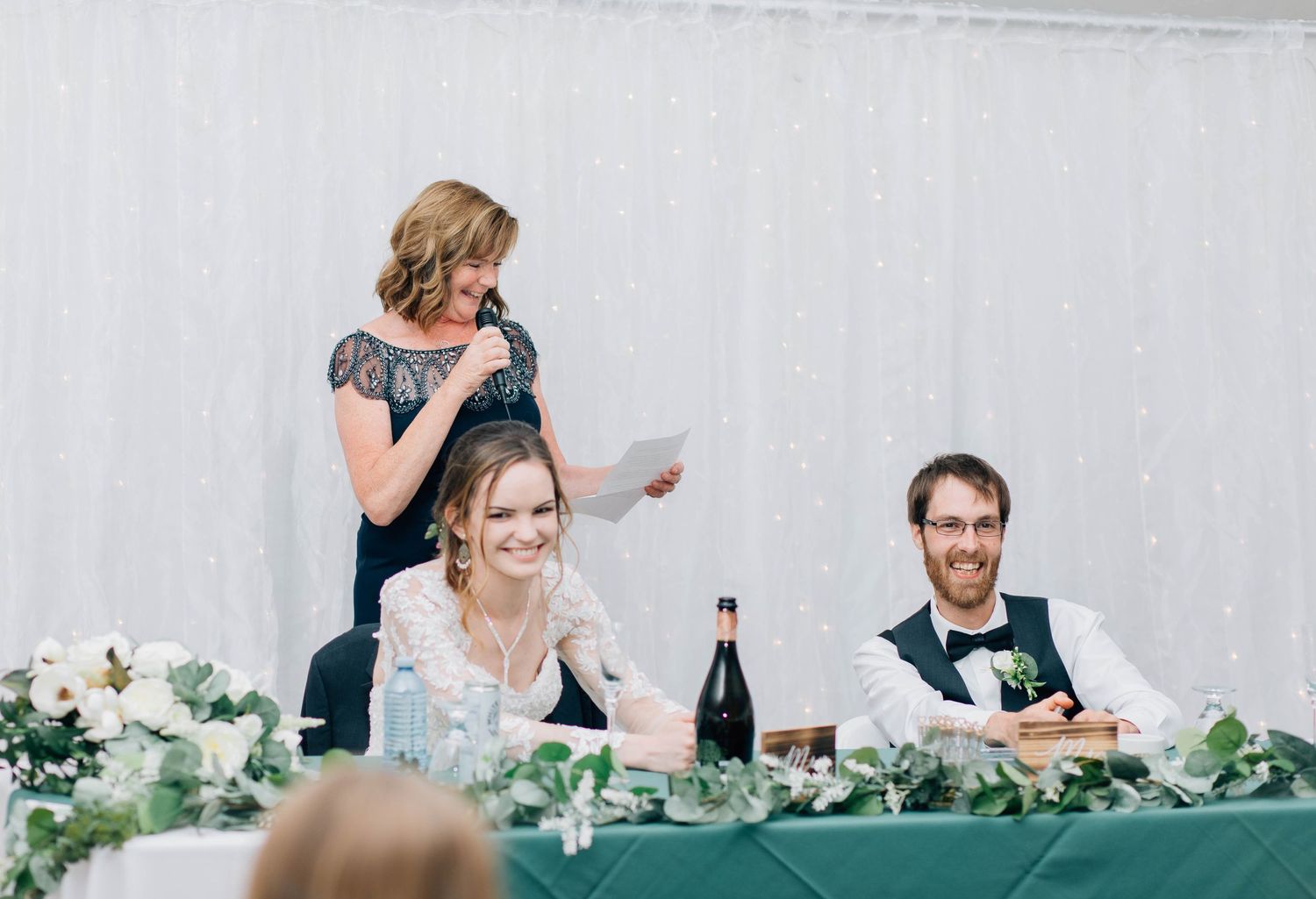 Woman reads touching speech at wedding reception as smiling bride and groom enjoy the moment.