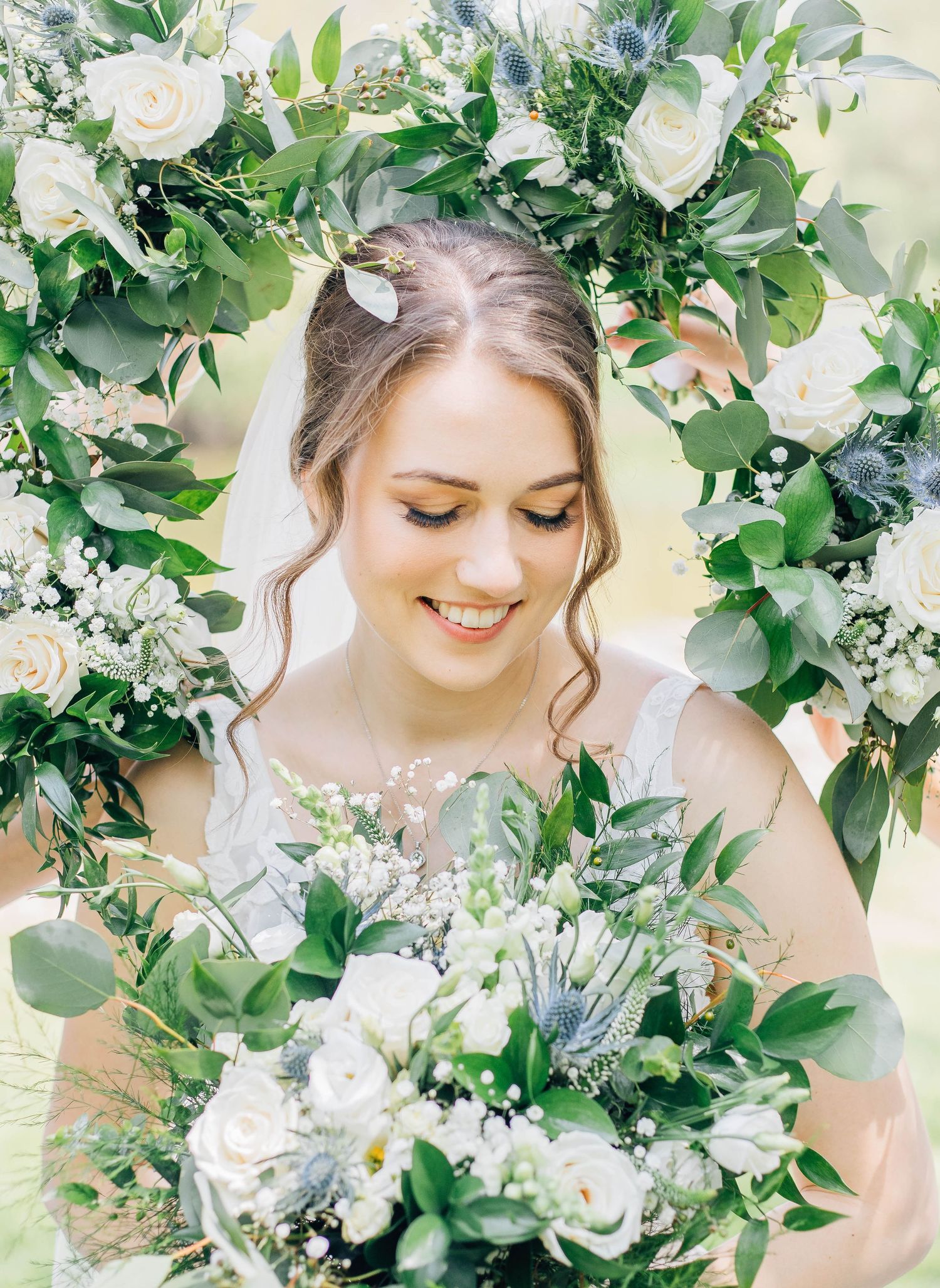 Smiling bride framed by a lush floral arch of white roses and greenery holding a matching bouquet.
