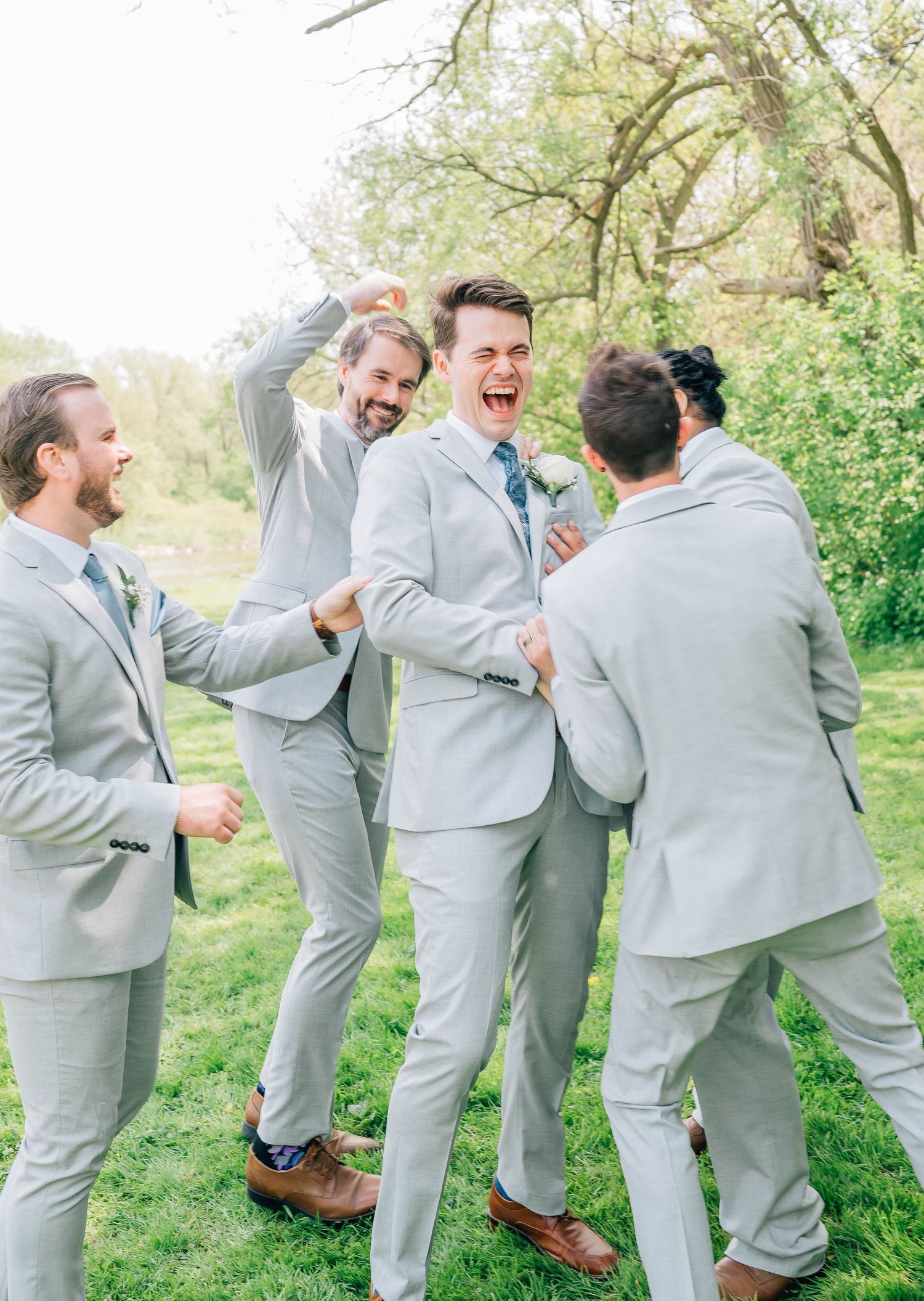 Groomsmen playfully celebrate with laughing groom in light gray suit on green lawn at spring wedding.