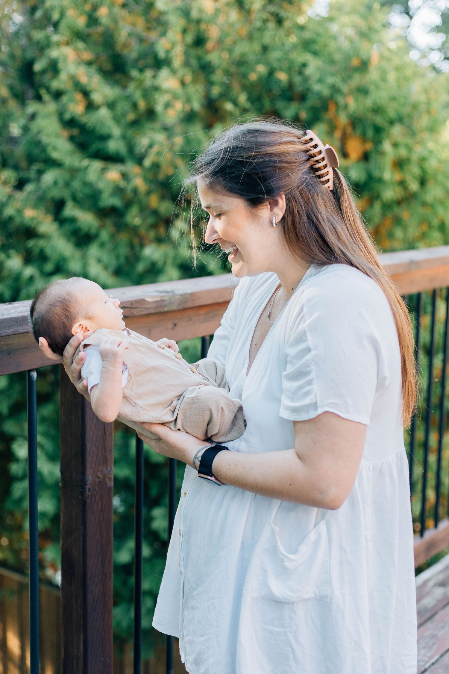 Smiling mother in white dress holding newborn baby on outdoor wooden deck.