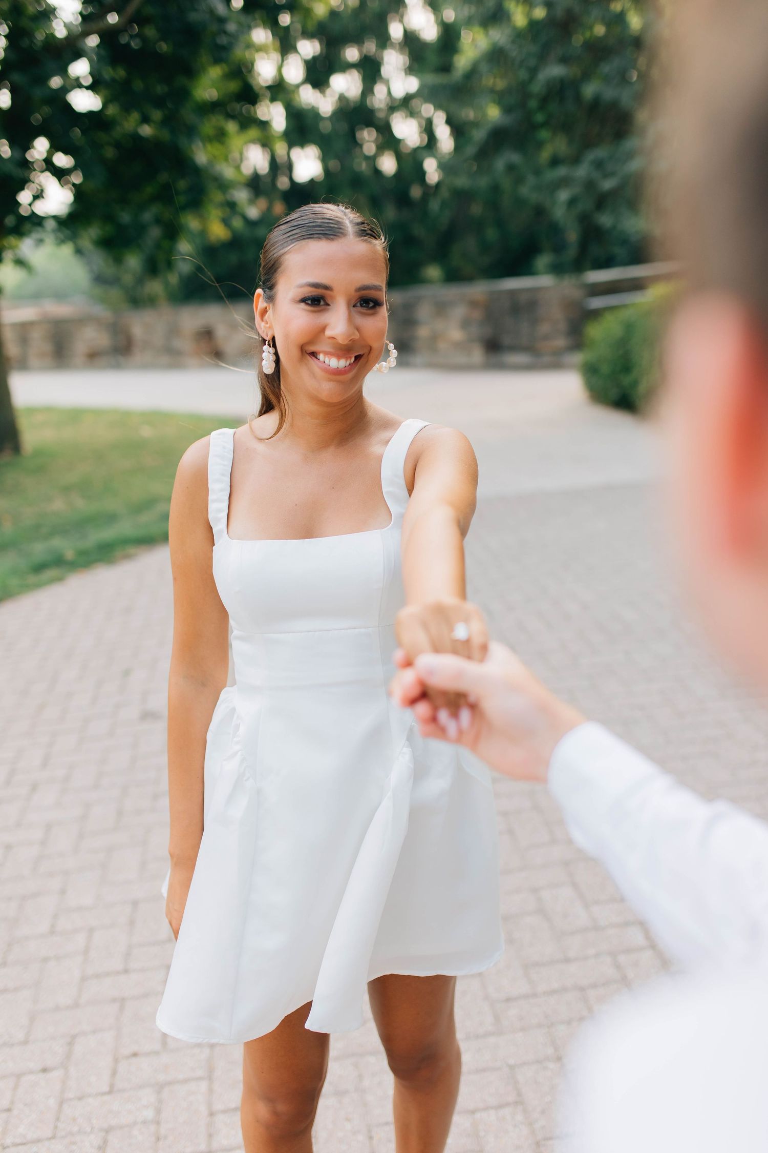 Smiling bride in white mini dress reaching out to hold her partners hand on a brick garden path in London Ontario