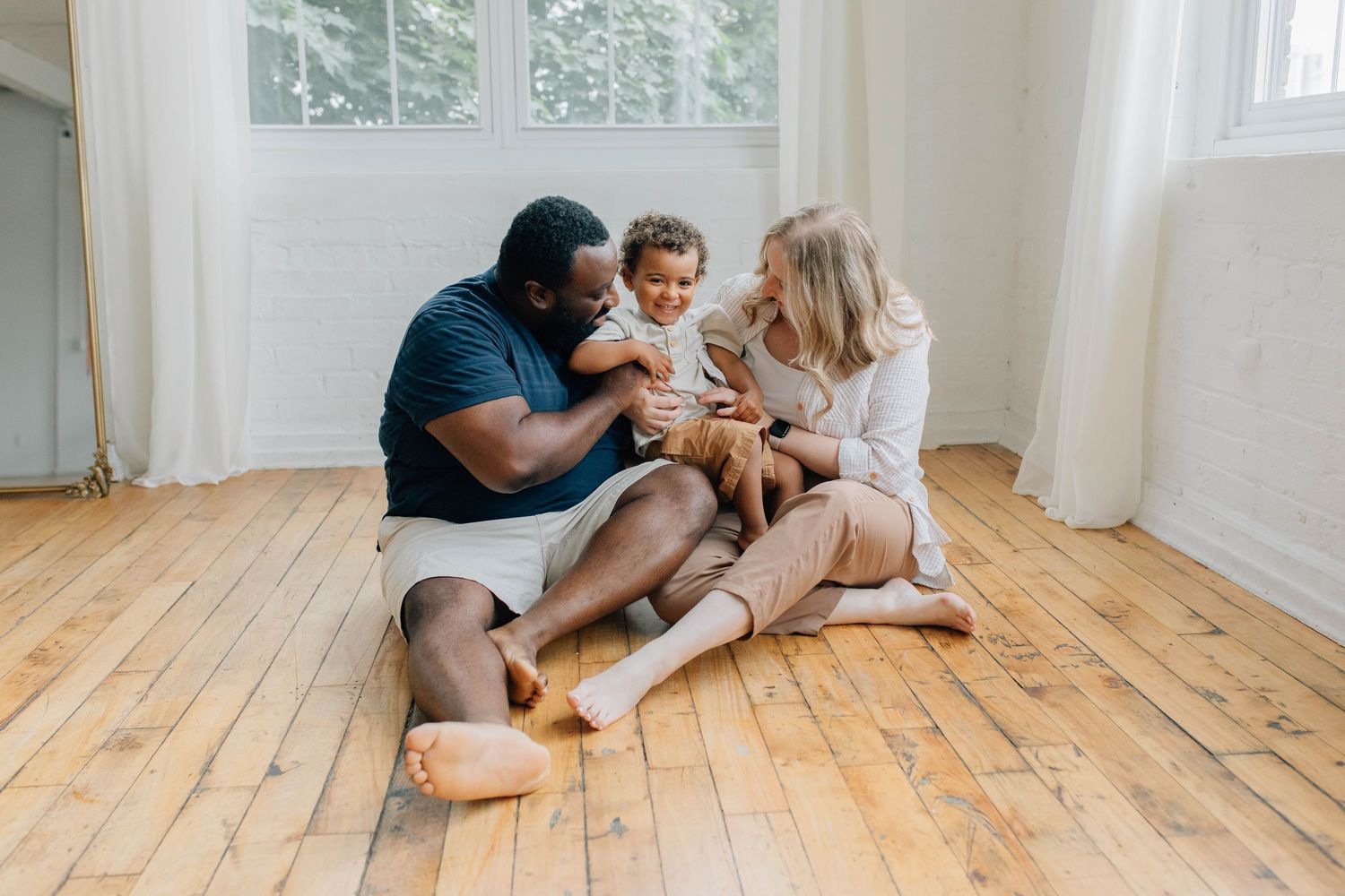 Interracial family sitting on hardwood floor, parents playing with happy toddler in bright white room.