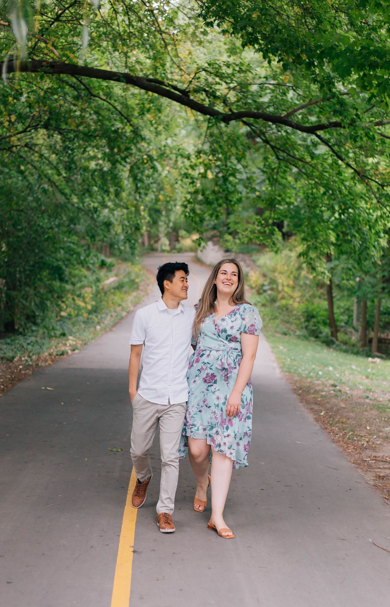 Couple walking hand in hand down a tree-lined path, smiling at each other.