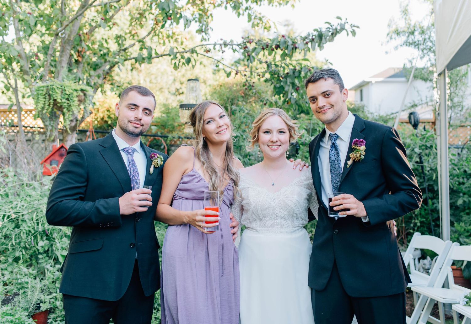 Four wedding guests pose together outdoors, two men in suits and two women in purple and white dresses.