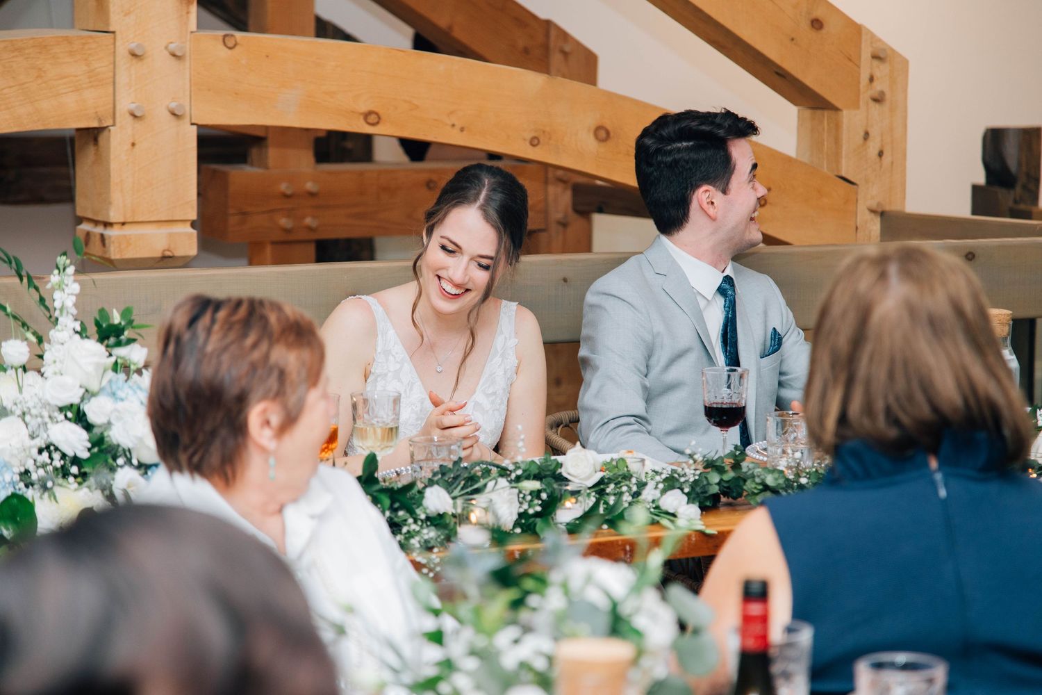 Smiling bride in white dress seated at floral wedding reception table with guests.